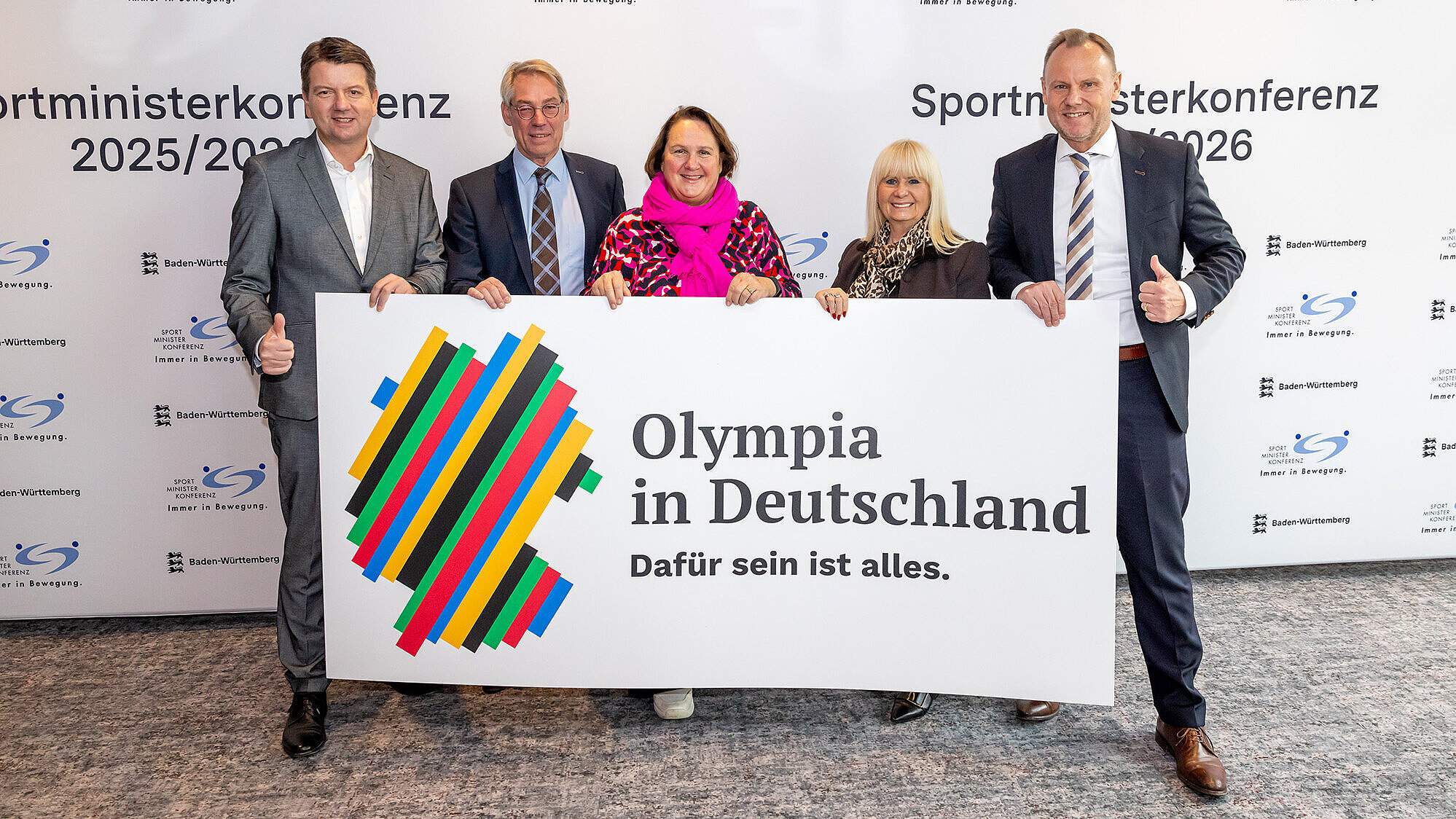 Auf dem Bild stehen von links nach rechts  Innen- und Sportstaatssekretär Sandro Kirchner, SMK-Vorsitzende Frau Schopper, Herr MDgt Schwank (Leiter Abteilung III, Sport und Ehrenamt Staatskanzlei NW), Frau Senatorin Spranger (BE) und Herrn Senator Grote (HH) mit einem Plakat in der Hand auf dem steht "Olympia in Deutschland".
