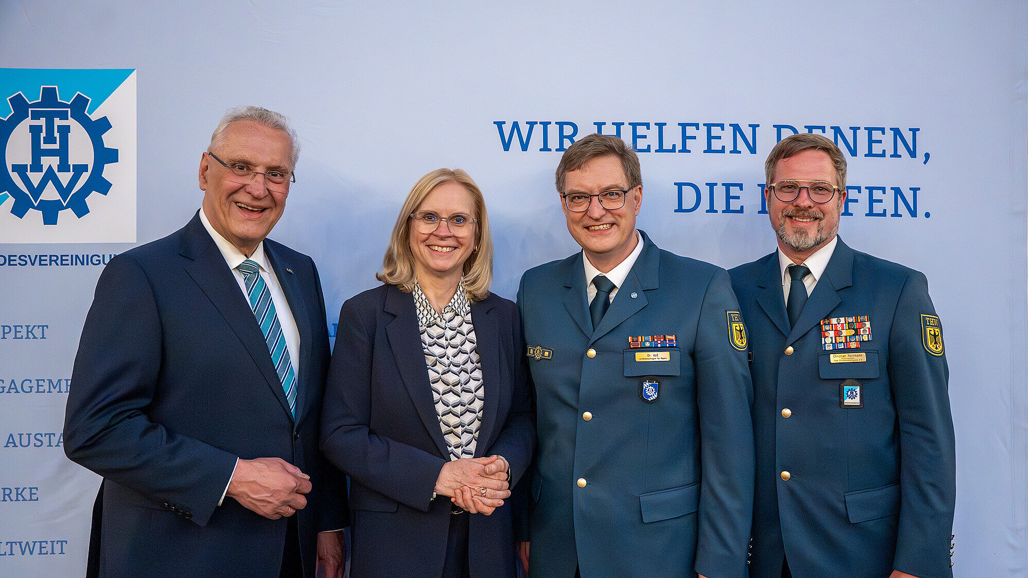 Vier Personen vor hellem Banner mit THW‑Logo; zwei Männer in THW‑Uniform, eine Frau und ein Mann in dunklem Anzug, Gruppenfoto.