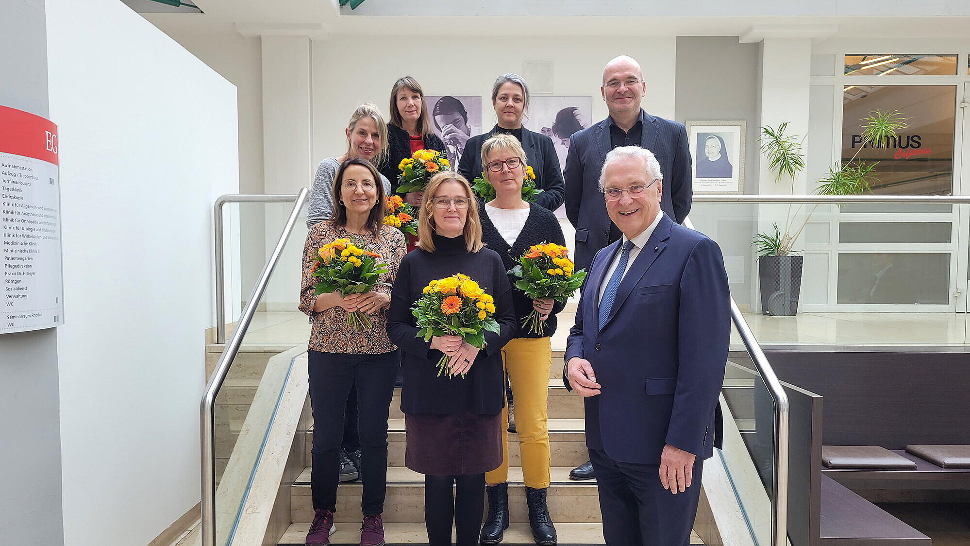 Eine Gruppe von acht Personen steht auf einer Treppe in einem hellen Innenraum. Sechs Personen halten jeweils einen Blumenstrauß mit gelben und orangen Blumen. Vorne rechts steht Innenminister Joachim Herrmann ein im dunklen Anzug mit Krawatte. Im Hintergrund sind Pflanzen und ein Schild mit der Aufschrift „Primus“ zu sehen. Die Treppe hat Edelstahlgeländer auf beiden Seiten.