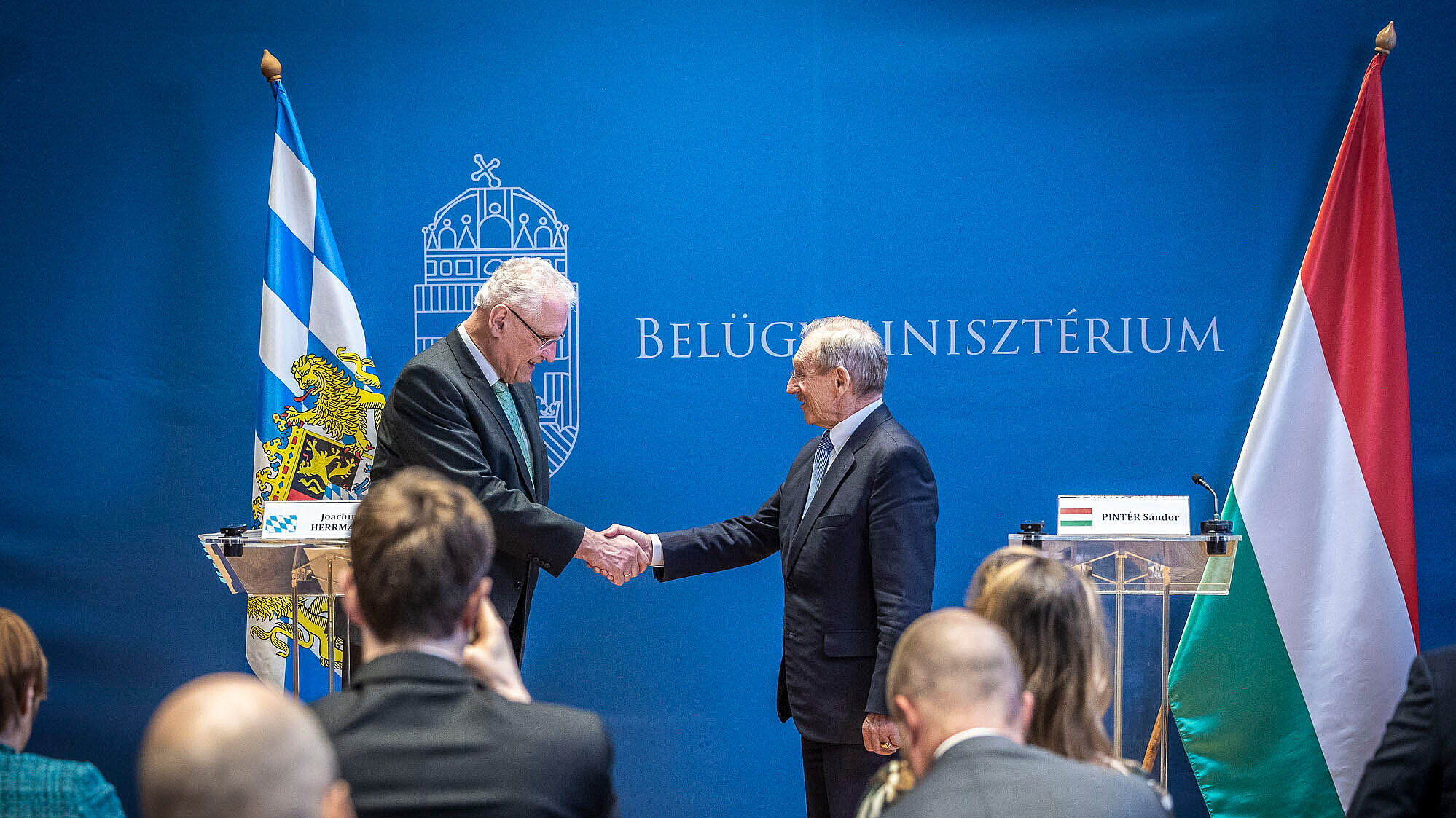 Innenraum bei einer offiziellen Presse- oder Gesprächsrunde im ungarischen Innenministerium. Personen: Innenminister Joachim Herrmann li. im Bild und ungarischen Innenminister Dr. Sandor Pinter in formeller Kleidung stehen an transparenten Rednerpulten; auf den Pulten sind Namensschilder, die hier nicht wiedergegeben werden.