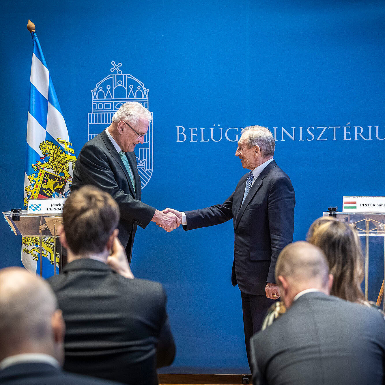 Innenraum bei einer offiziellen Presse- oder Gesprächsrunde im ungarischen Innenministerium. Personen: Innenminister Joachim Herrmann li. im Bild und ungarischen Innenminister Dr. Sandor Pinter in formeller Kleidung stehen an transparenten Rednerpulten; auf den Pulten sind Namensschilder, die hier nicht wiedergegeben werden.