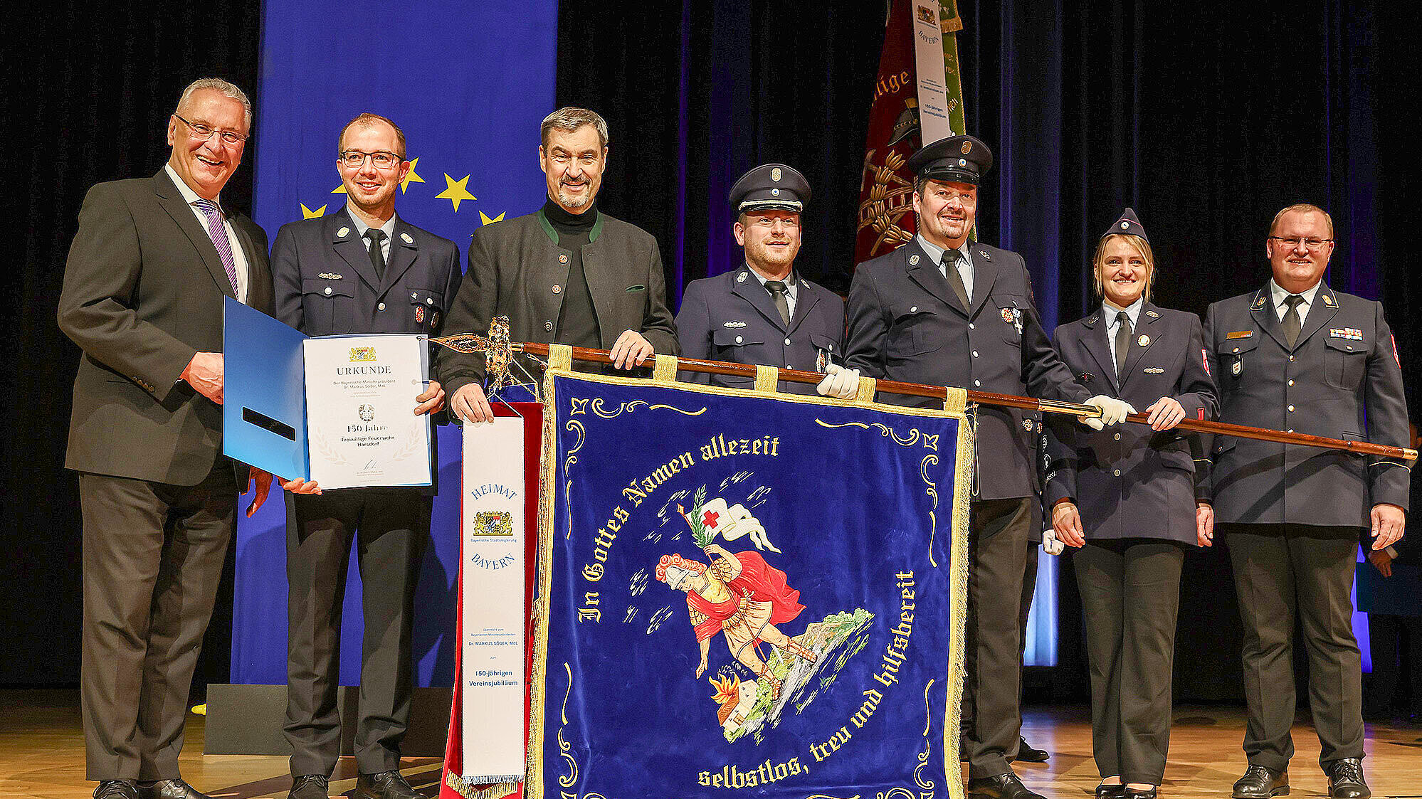 Eine Gruppe von sieben Personen (1. von li. nach rechts Innenminister Joachim Herrmann, 3.v. li. MPr Dr. Markus Söder)steht auf einer Bühne vor einem blauen Vorhang mit gelben Sternen. Einige der Personen tragen Uniformen der Feuerwehr, andere formelle Kleidung. Eine Person hält eine Urkunde, eine andere eine große blaue Fahne mit der Aufschrift „In Gottes Namen allezeit selbstlos, treu und hilfsbereit“ und einem Bild eines Feuerwehrmanns mit Helm und Axt. Die Fahne hat einen roten Rand und eine weiße Banderole mit der Aufschrift „Heimat Bayern“ und „150-jähriges Vereinsjubiläum“.
