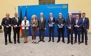 alle Preisträger der Verleihung mit Innenminister Joachim Herrmann und Staatssekretär Sandro Kirchner vor einer blauen Pressewand im Odeon des StMI