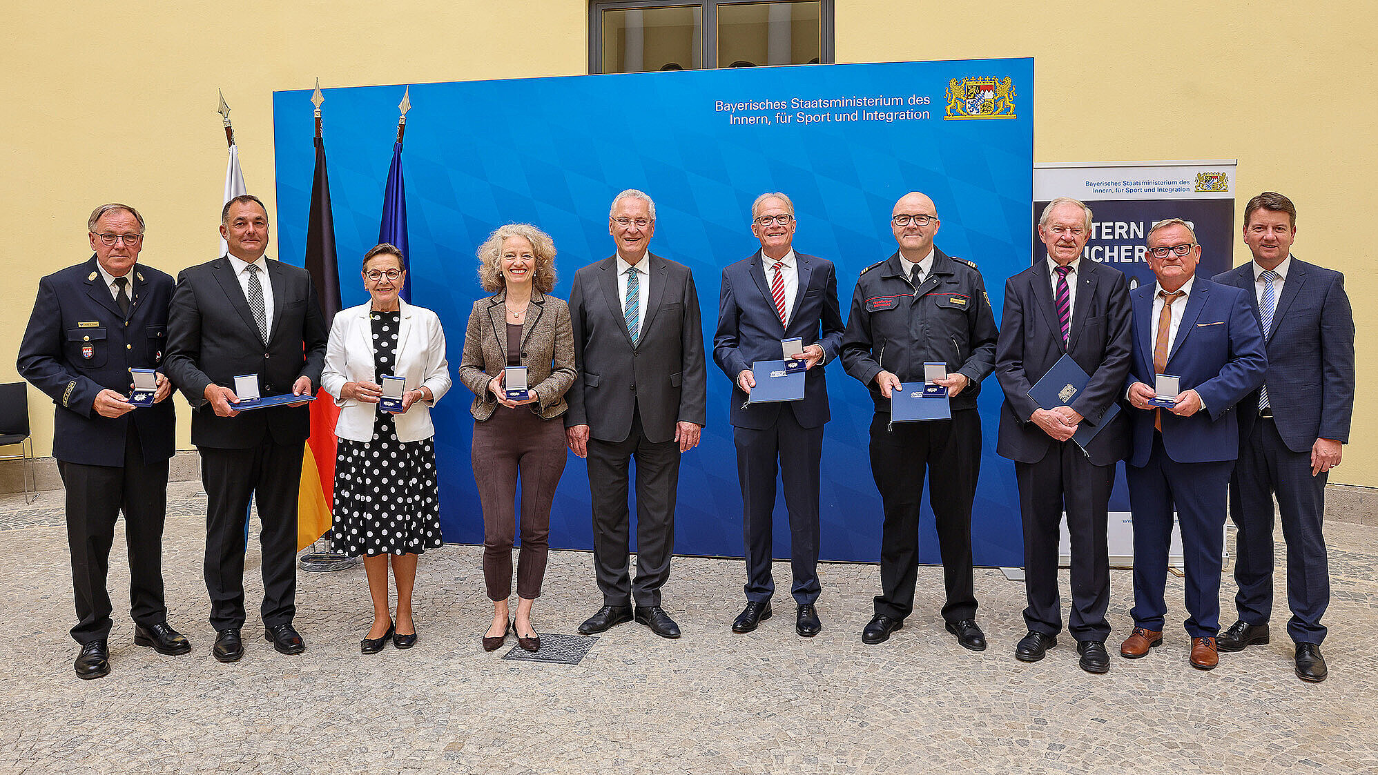 alle Preisträger der Verleihung mit Innenminister Joachim Herrmann und Staatssekretär Sandro Kirchner vor einer blauen Pressewand im Odeon des StMI