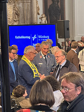 Menschen in einem Kirchenraum während einer Veranstaltung; im Zentrum mehrere Personen im Gespräch, links ein großer blauer Bildschirm mit der Aufschrift „Katholikentag Würzburg“, im Vordergrund unscharfe Zuhörer.