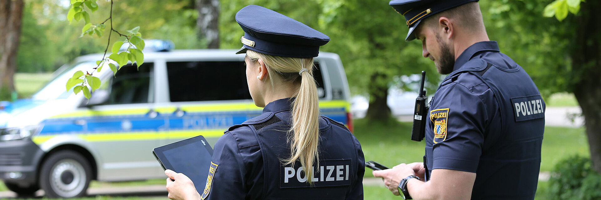 Polizist und Polizistin von hinten im Streifendienst, die auf Mobile Geräte schauen und im Hintergrund ein Polizeiauto auf einer Wiese