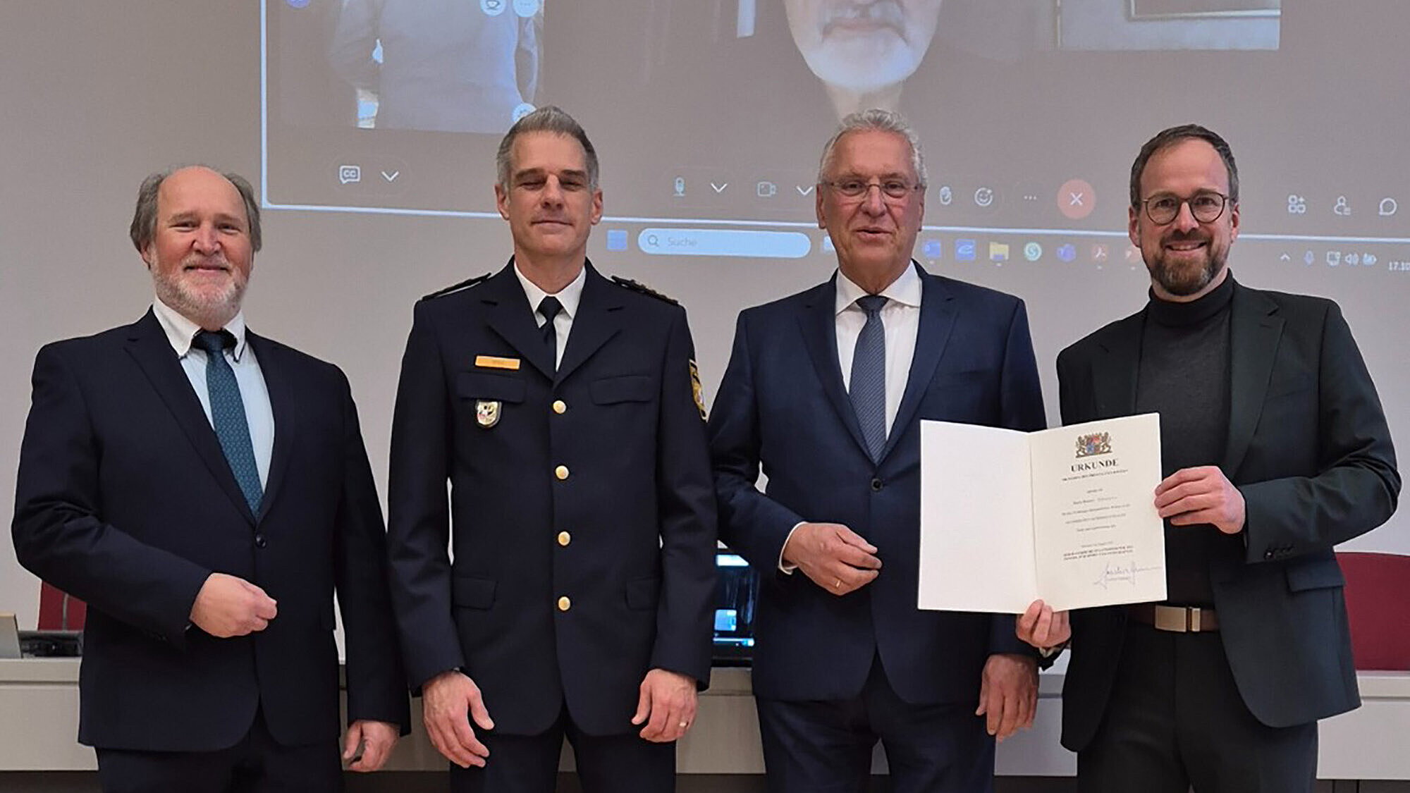Vier Männer (2. v. li. Innenminister Joachim Herrmann) in formeller Kleidung, darunter ein Mann in Uniform, die bei einer Zeremonie eine Urkunde präsentieren.