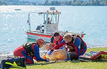 Drei Rettungskräfte an Land mit einer Gummipuppen zum Simulieren einer Rettungssituation, dahinter ein Rettungsboot auf dem Wasser