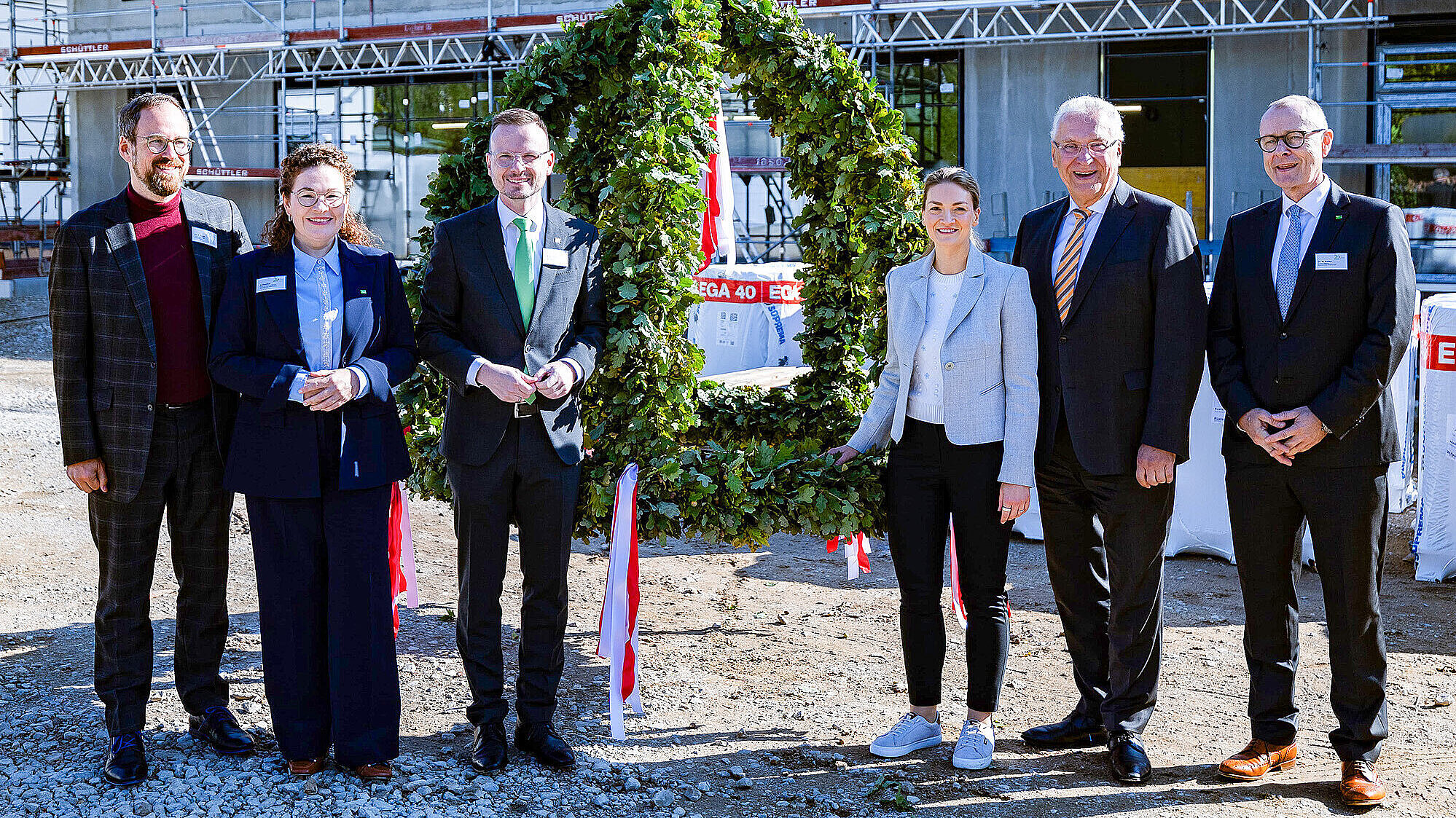 Auf dem Bild sind sechs Personen (zweite von links Innenminister Joachim Herrmann, dritte von links Staatsministerin Judith Gerlach) bei einem Festakt vor einer Baustelle zu sehen. Sie stehen nebeneinander, vor einem grünen Kranz, der für die Zeremonie aufgestellt wurde. Die Personen tragen formelle Kleidung, darunter Anzüge und Blazer. Im Hintergrund sind Baugerüste und Bauarbeiten sichtbar, was auf eine Baustelle hinweist.
