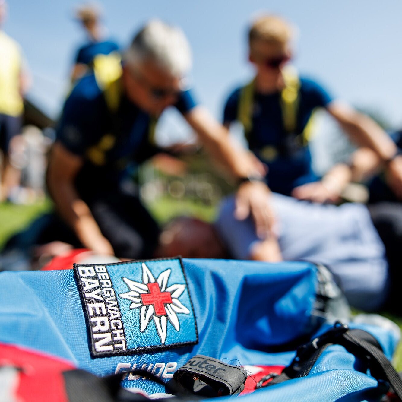 Auf dem Foto ist im Vordergrund ein Rucksack mit dem Logo der Bergwacht Bayern zu sehen und im Hintergrund sind zwei Bergwacht-Mitglieder bei Erste-Hilfe-Übungen verschwommen erkennbar.