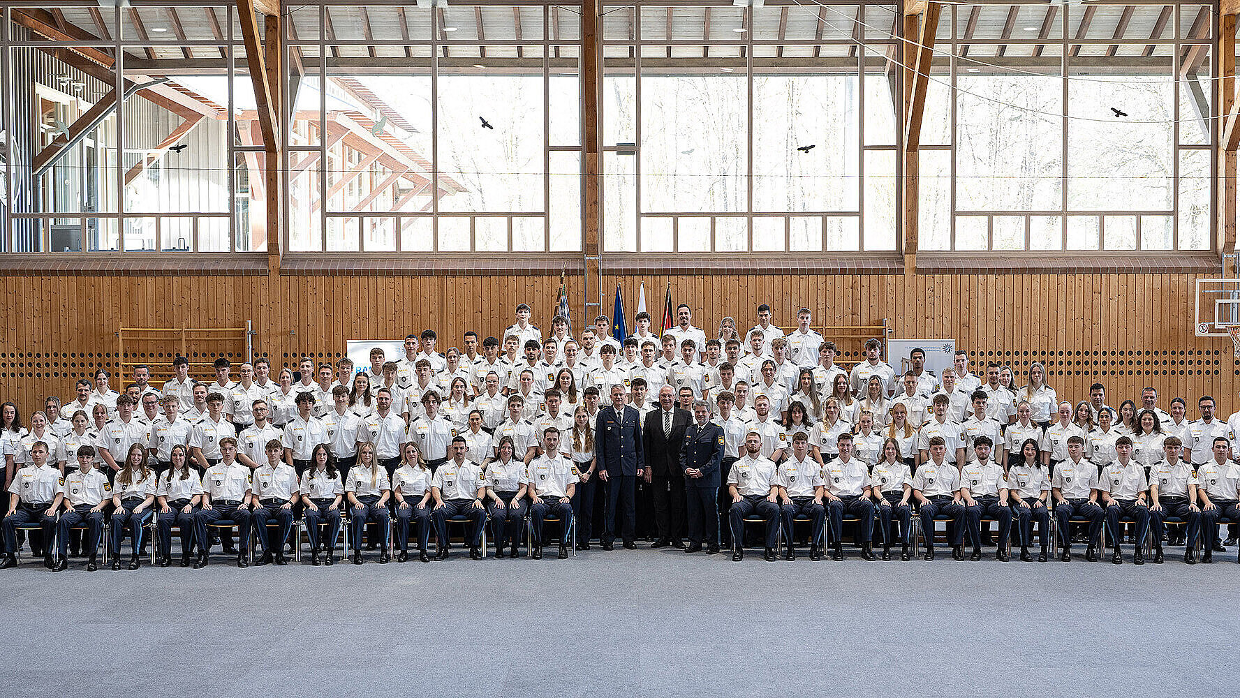 Große Gruppe uniformierter Personen in einer Halle vor Holzverkleidung und großen Fenstern.