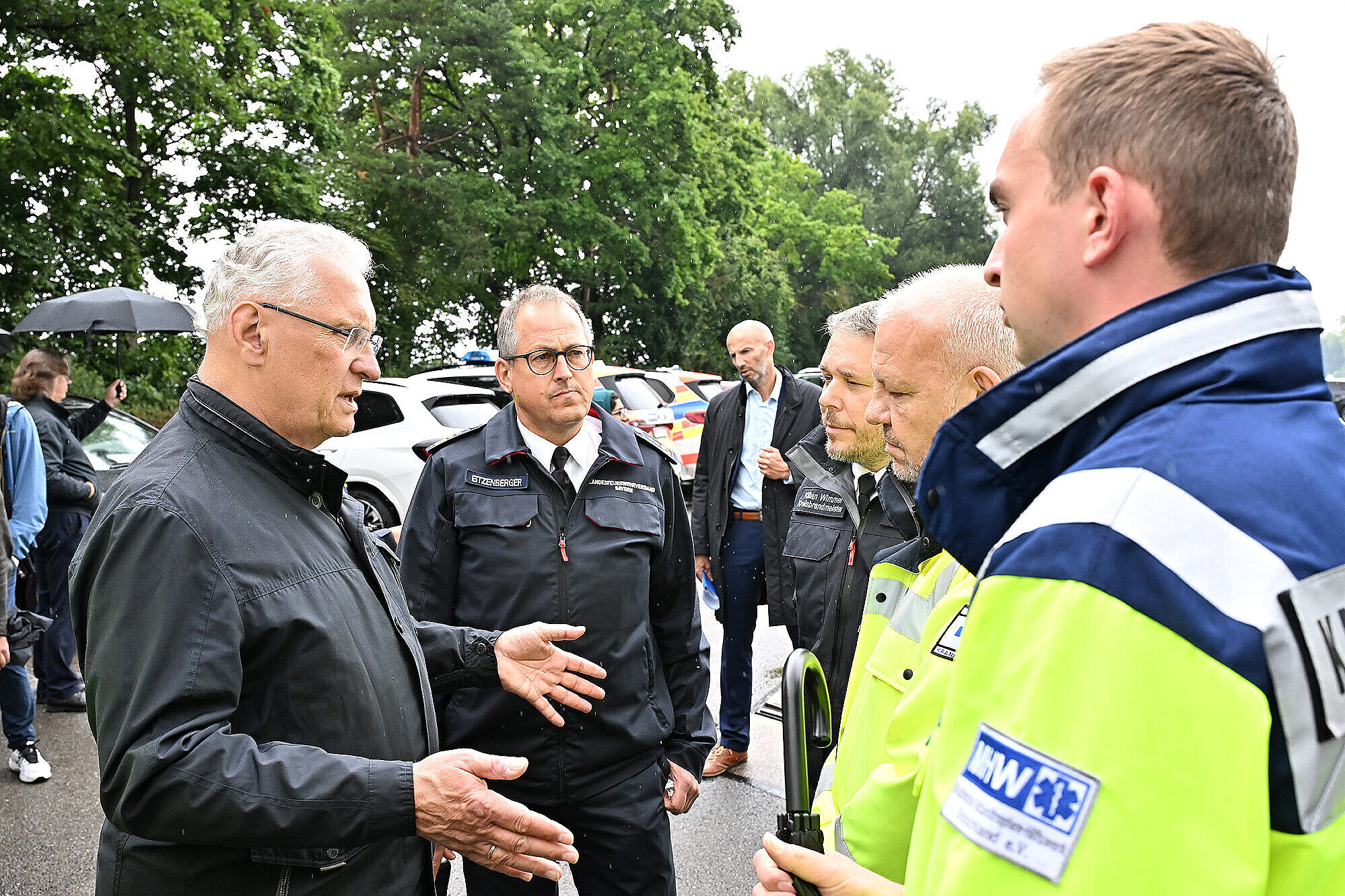 Bayerns Innenminister Joachim Herrmann im Gespräch mit Einsatzkräften bei einer Katastrophenschutzübung im Landkreis München. Im Bild zu sehen sind mehrere Männer in Einsatzkleidung, darunter Vertreter des Technischen Hilfswerks (THW) und der Katastrophenschutzbehörden. Die Szene spielt sich im Freien bei regnerischem Wetter ab. Im Hintergrund stehen Einsatzfahrzeuge und weitere Personen.