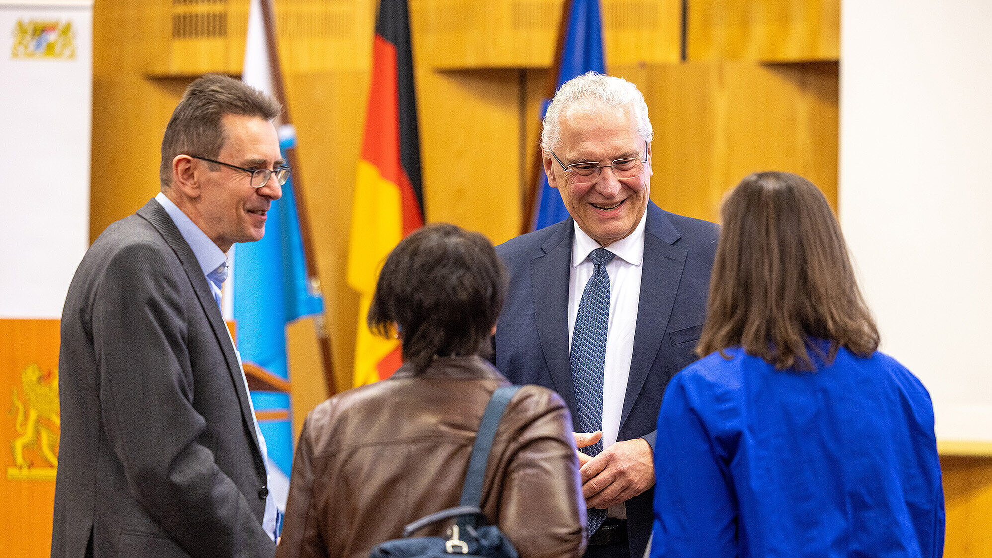Vier Personen stehen in einem Raum mit hölzernen Wänden und diskutieren miteinander. Im Hintergrund sind mehrere Fahnen zu sehen, darunter die deutsche Flagge. Zwei Personen sind von hinten zu sehen, eine trägt eine braune Lederjacke und hat eine Tasche über der Schulter, die andere trägt eine blaue Jacke. Die beiden anderen Personen tragen Anzüge (rechts steht Innenminister Joachim Herrmann)..