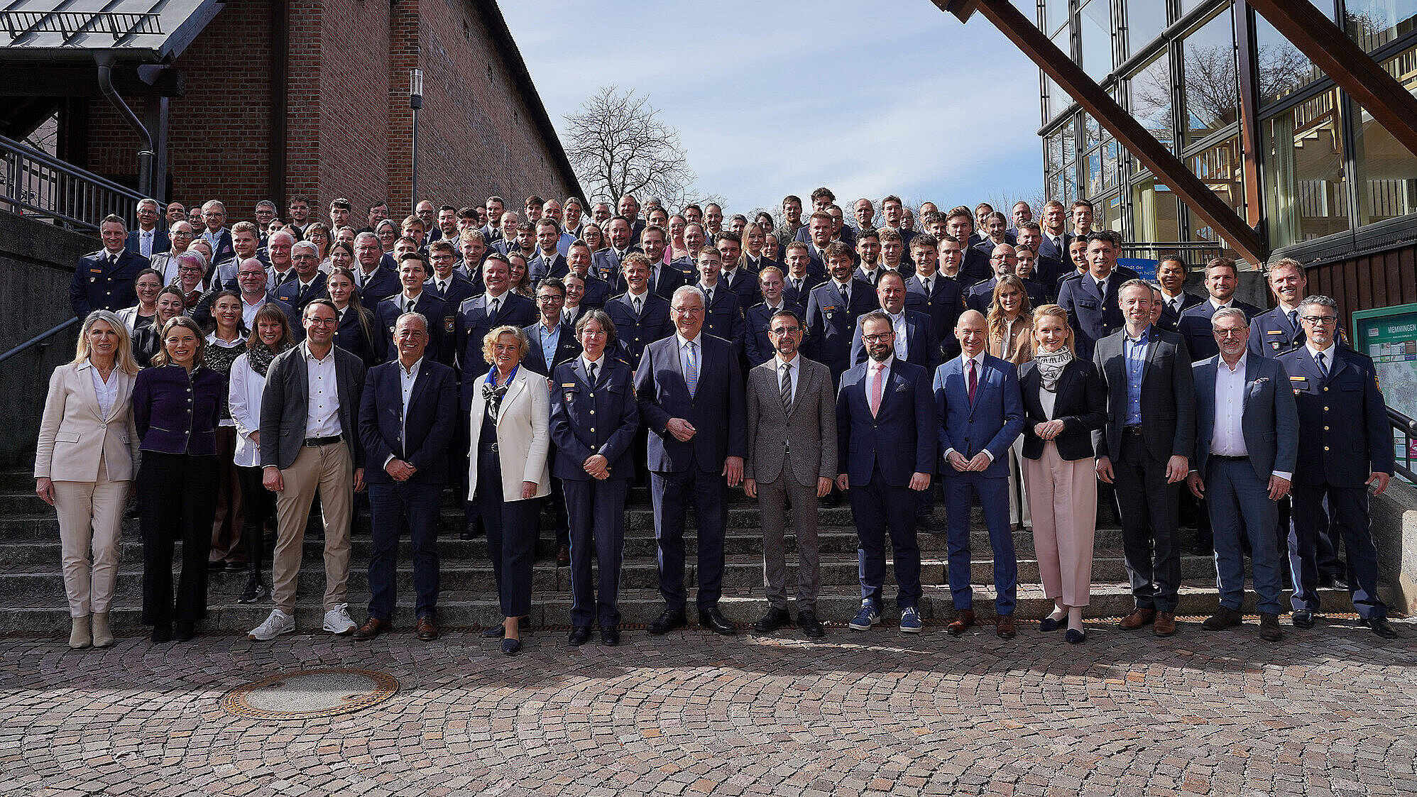 Gruppenfoto mehrerer Dutzend Mitarbeiterinnen und Mitarbeiter, teils in Uniform, teils in ziviler Kleidung, versammelt auf einer Treppe vor einem Verwaltungsgebäude bei klarem Himmel.