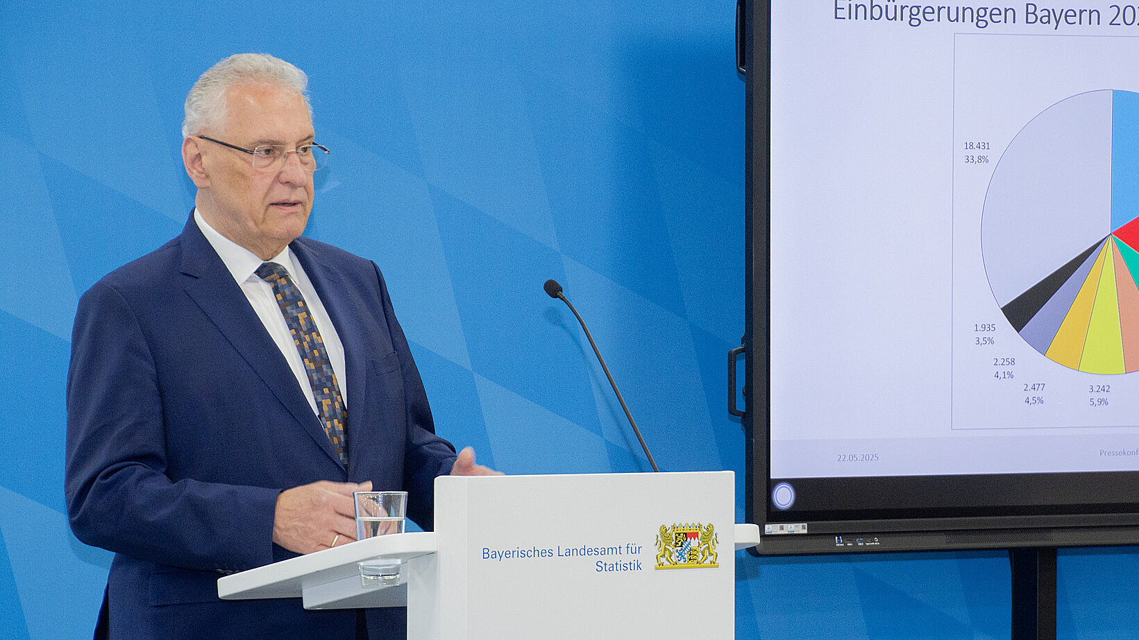 Innenminister Herrmann am Rednerpult vor blauer Pressewand im Hintergrund Bildschirm mit abgebildeter Statistik
