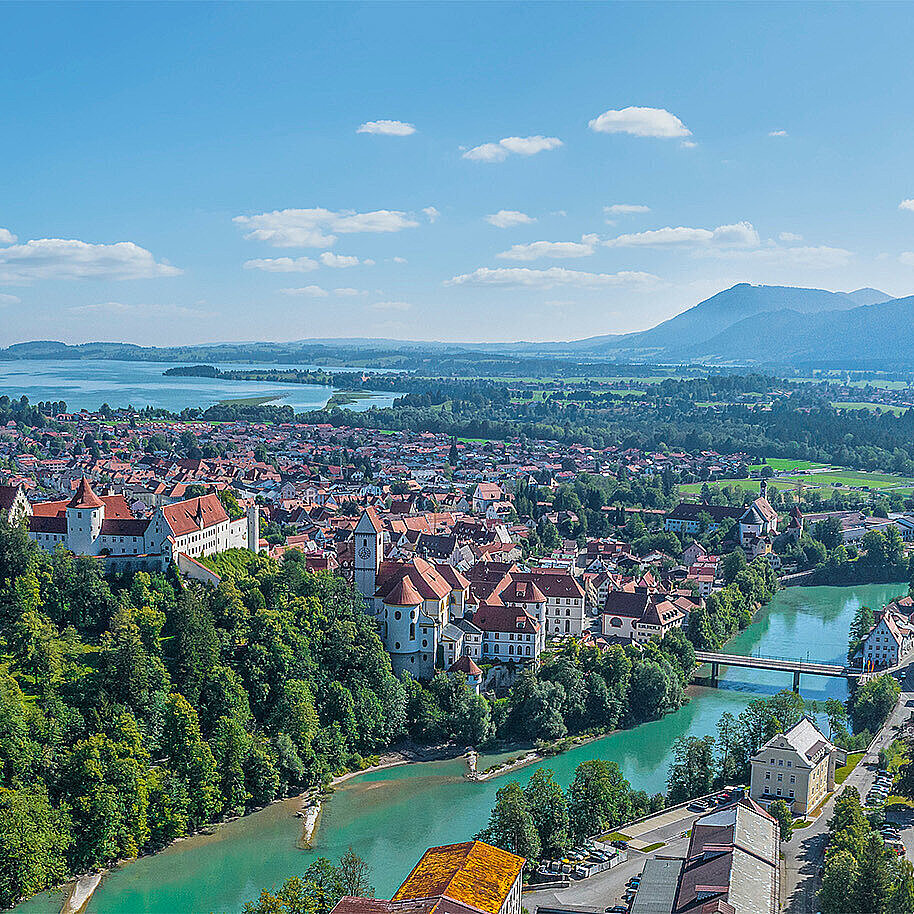 Auf dem Bild ist die Stadt Füssen aus zu sehen, im Vordergrund ein Fluss, im Hintergrund die Berge.