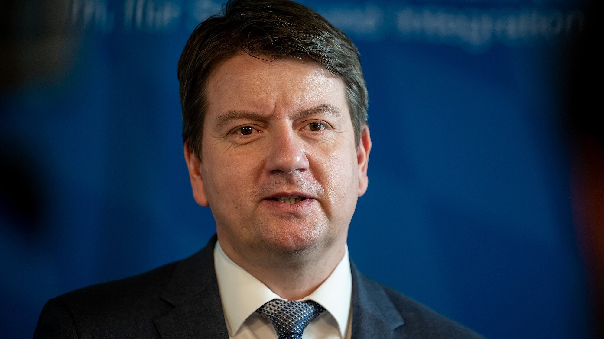Mann im dunklen Anzug vor blauem Hintergrund; weißes Hemd und gemusterte blaue Krawatte; Gesicht durch quadratisches Feld unkenntlich.