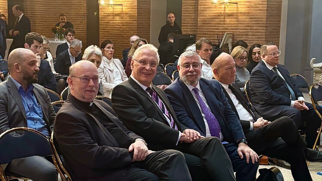 Eine Gruppe von Personen sitzt in einem Saal auf Reihen von blauen Stühlen (zweite von li Innenminister Joachim Herrmann)mit goldfarbenen Gestellen. Die meisten tragen formelle Kleidung, darunter Anzüge und Hemden. Im Hintergrund sind weitere Teilnehmer zu sehen, die ebenfalls sitzen oder stehen. Die Wände sind mit hellem Backstein verkleidet, und es gibt Wandlampen, die warmes Licht spenden. Der Boden ist mit einem gemusterten Mosaik ausgelegt.