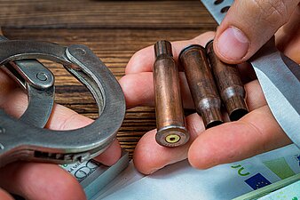 Hände halten Munition, ein Messer und Handschellen, darunter liegen Geldscheine