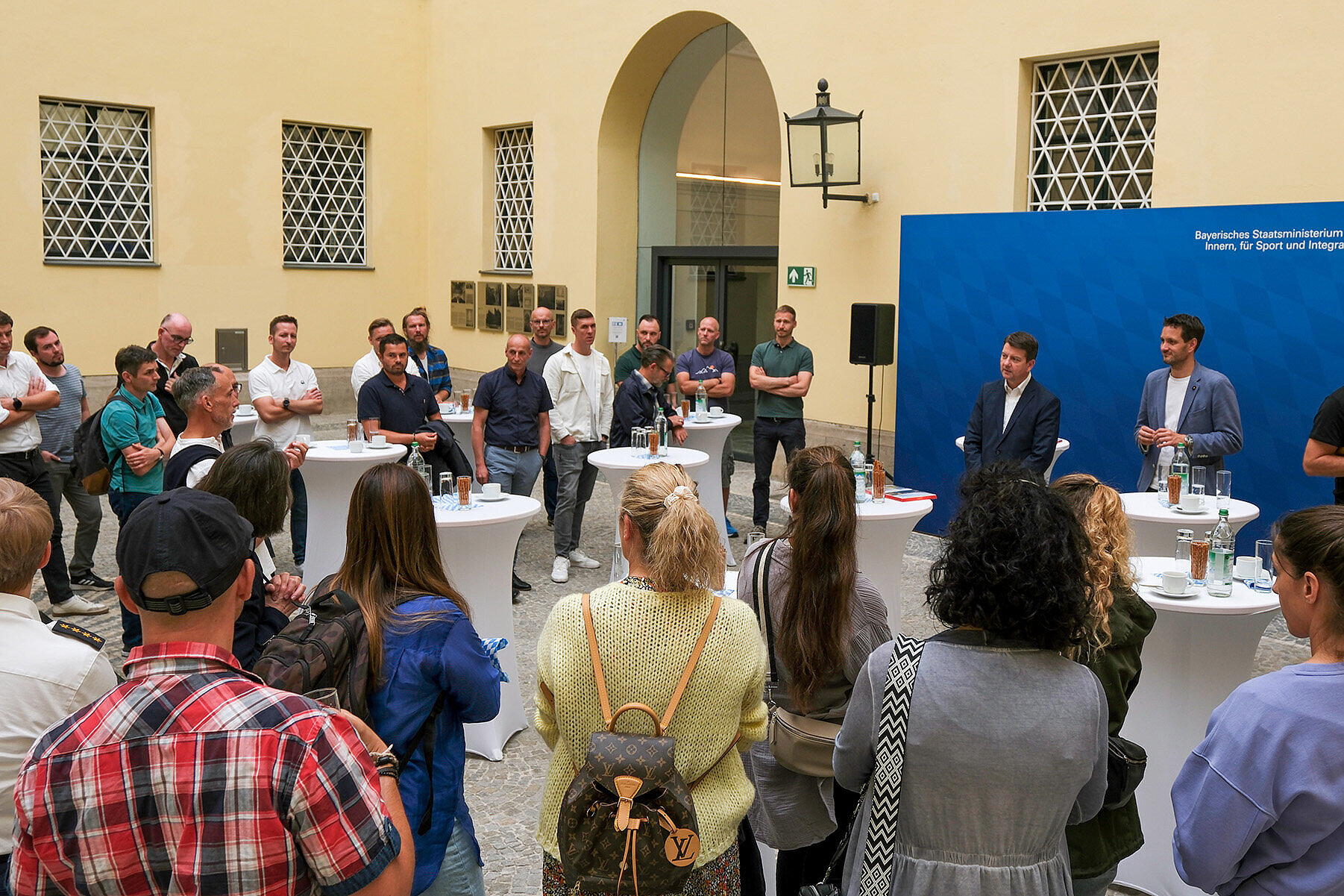 Staatssekretär Sandro Kirchner steht im Innenhof des Bayerischen Innenministeriums vor einer blauen Pressewand und spricht zu einer Gruppe von Polizistinnen und Polizisten. Um ihn versammeln sich mehrere Personen an Stehtischen, die aufmerksam zuhören. Im Hintergrund ist das Gebäude mit gelber Fassade und zwei vergitterten Fenstern zu sehen.