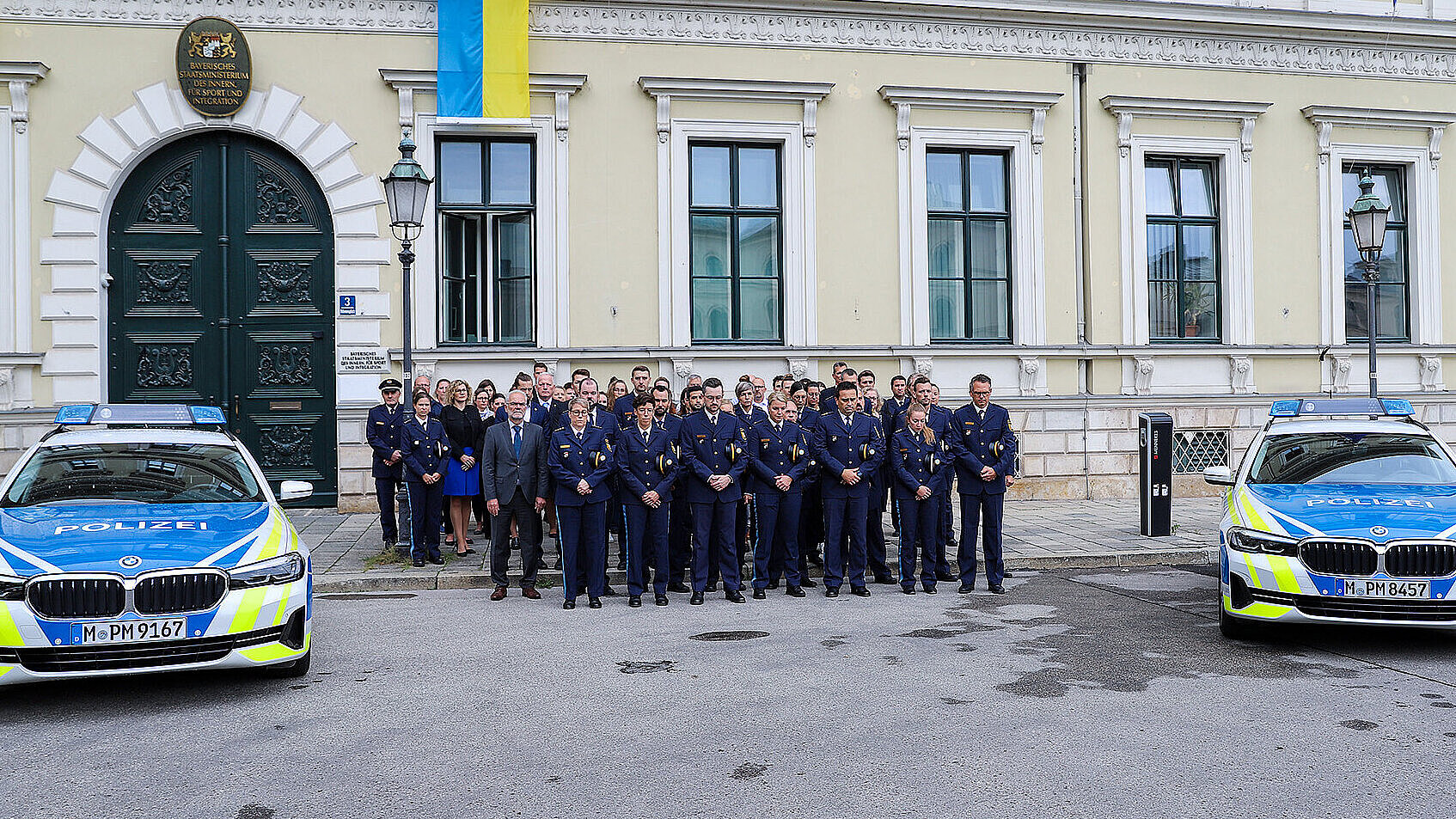 Polizisten stehen in einer Gruppe zwischen zwei Streifenwagen vor dem Eingang des Bayerischen Innenministeriums und halten eine Schweigeminute.