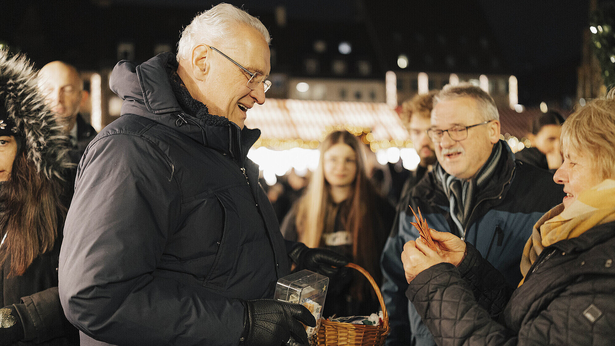 Innenminister Joachim Herrmann steht mit einer Spendenbox und den Sternstunden-Stern auf dem Christkindlesmarkt, vor ihm eine Ehepaar, dass einen Stern erwirbt.