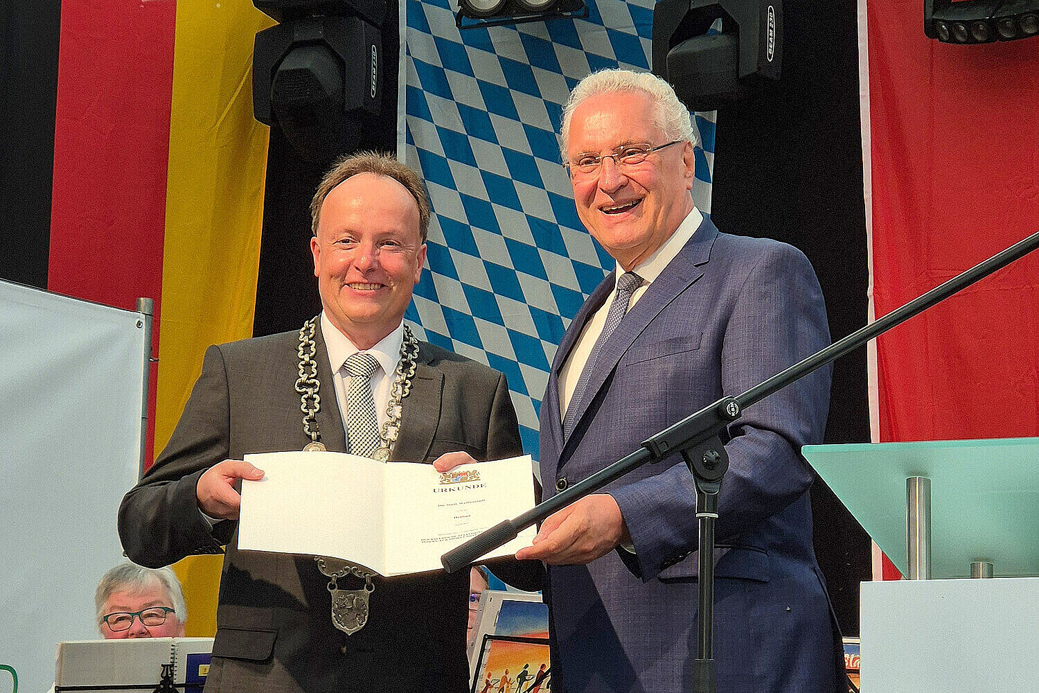 Innenminister Herrmann mit dem Bürgermeister Matthias Beck auf der Bühne in der Hand halten beide die Prädikatsurkunde, die Bad Weißenstadt zum Heilbad erklärt.