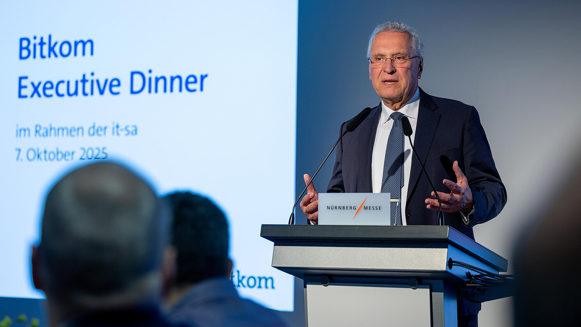 Innenminister Joachim Herrmann im Anzug am Rednerpult bei seiner Rede mit Blick ins Publikum, im Hintergrund sieht  man einen Screen mit der Aufschrift Bitcom