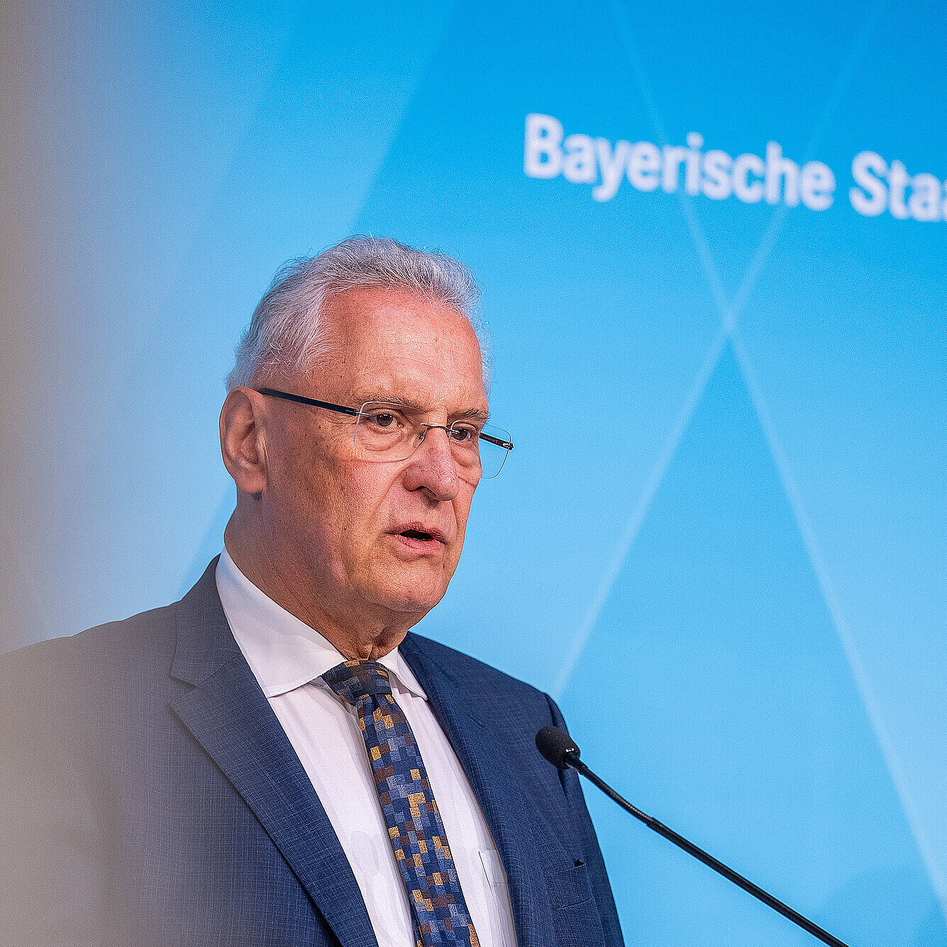 Mann in dunkelblauem Anzug und gemusterter Krawatte spricht an einem Rednerpult vor blauem Hintergrund.