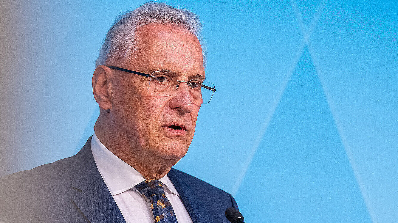 Mann in dunkelblauem Anzug und gemusterter Krawatte spricht an einem Rednerpult vor blauem Hintergrund.