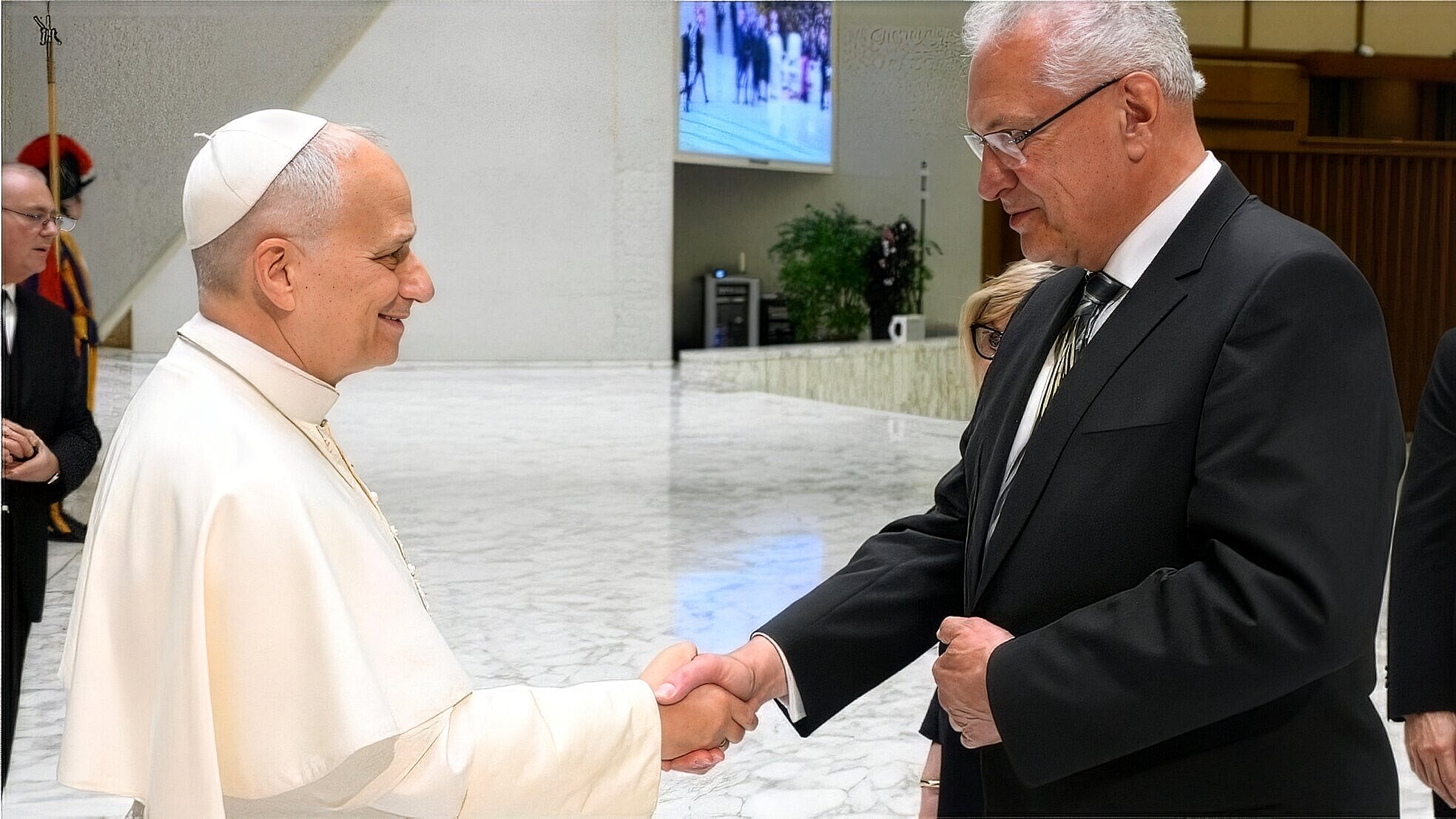 Zwei Personen schütteln sich die Hand: eine in weißem liturgischen Gewand mit Kappe, die andere in dunklem Anzug, in hellem Marmorsaal.