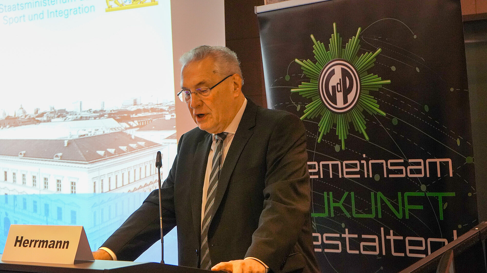 Innenminister Joachim Herrmann steht an einem Rednerpult mit einem Namensschild „Herrmann“. Im Hintergrund ist eine Projektionsfläche mit einem Bild eines historischen Gebäudes und ein Banner mit dem Logo der Gewerkschaft der Polizei (GdP) sowie dem Schriftzug „gemeinsam Zukunft gestalten“ zu sehen.