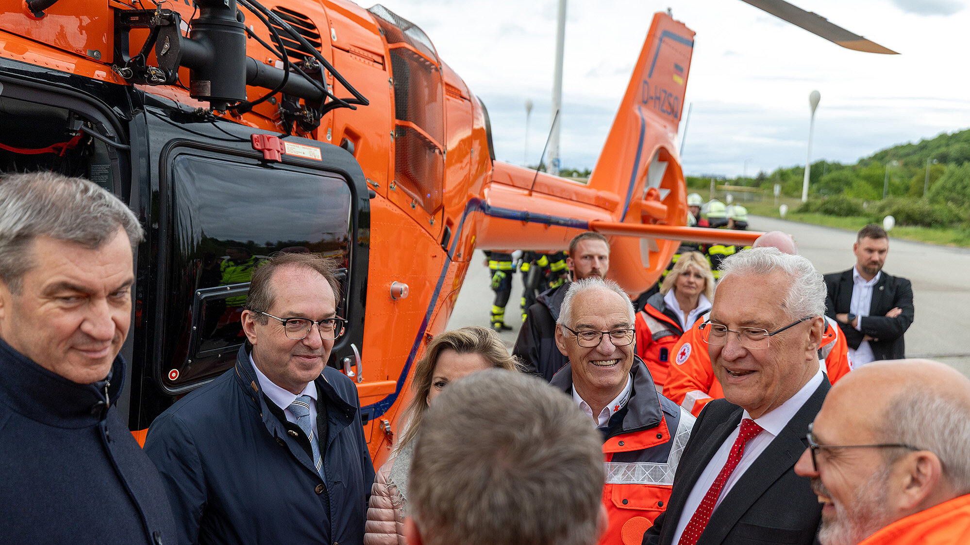 Personengruppe in Rettungsjacken und Anzügen steht neben einem orangefarbenen Rettungshubschrauber auf einem Flugfeld bei bewölktem Himmel
