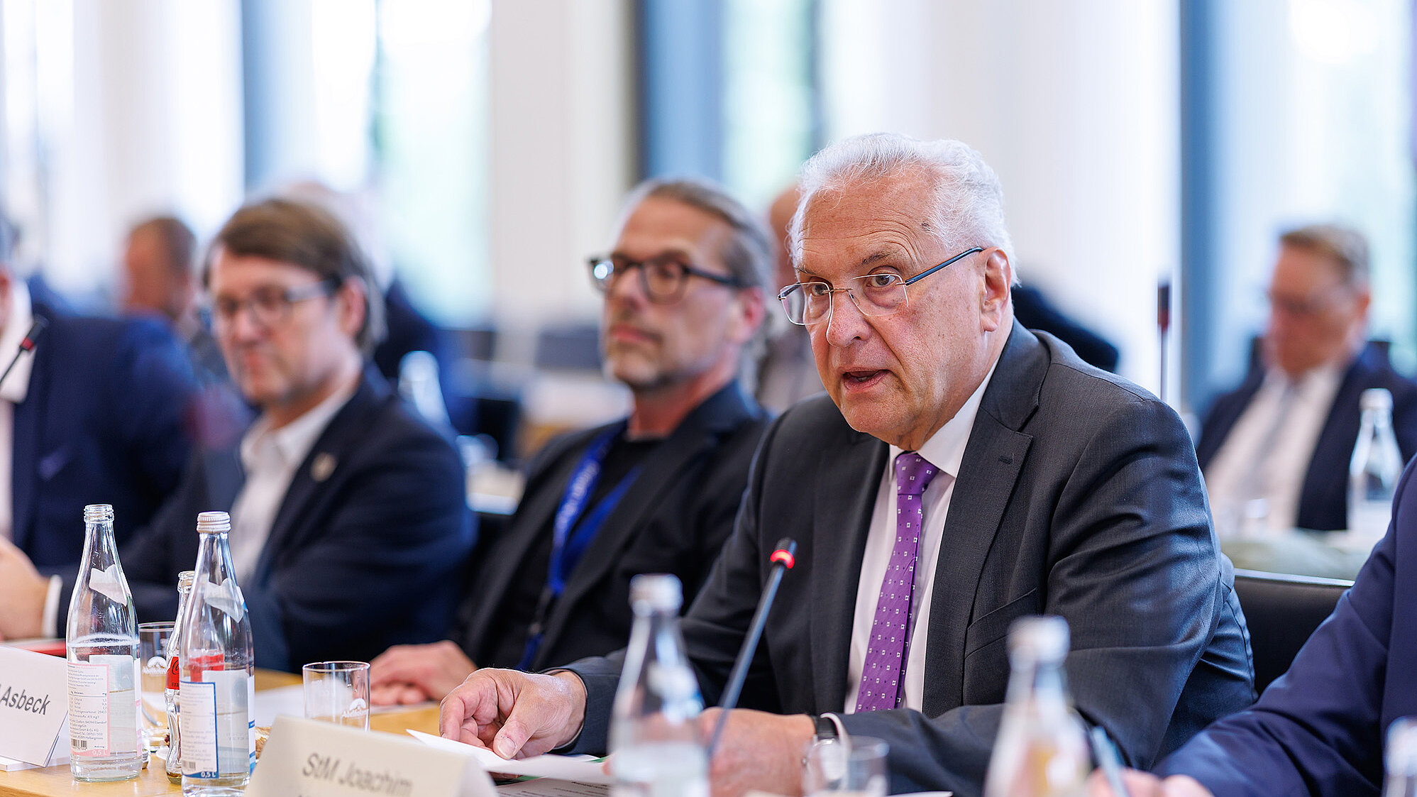 Konferenzraum: mehrere Personen in Anzügen sitzen an einem Tisch mit Mikrofonen, Wasserflaschen und Unterlagen; ein Mann im Vordergrund mit dunklem Anzug und violetter Krawatte spricht ins Mikrofon. Gesichter unscharf.