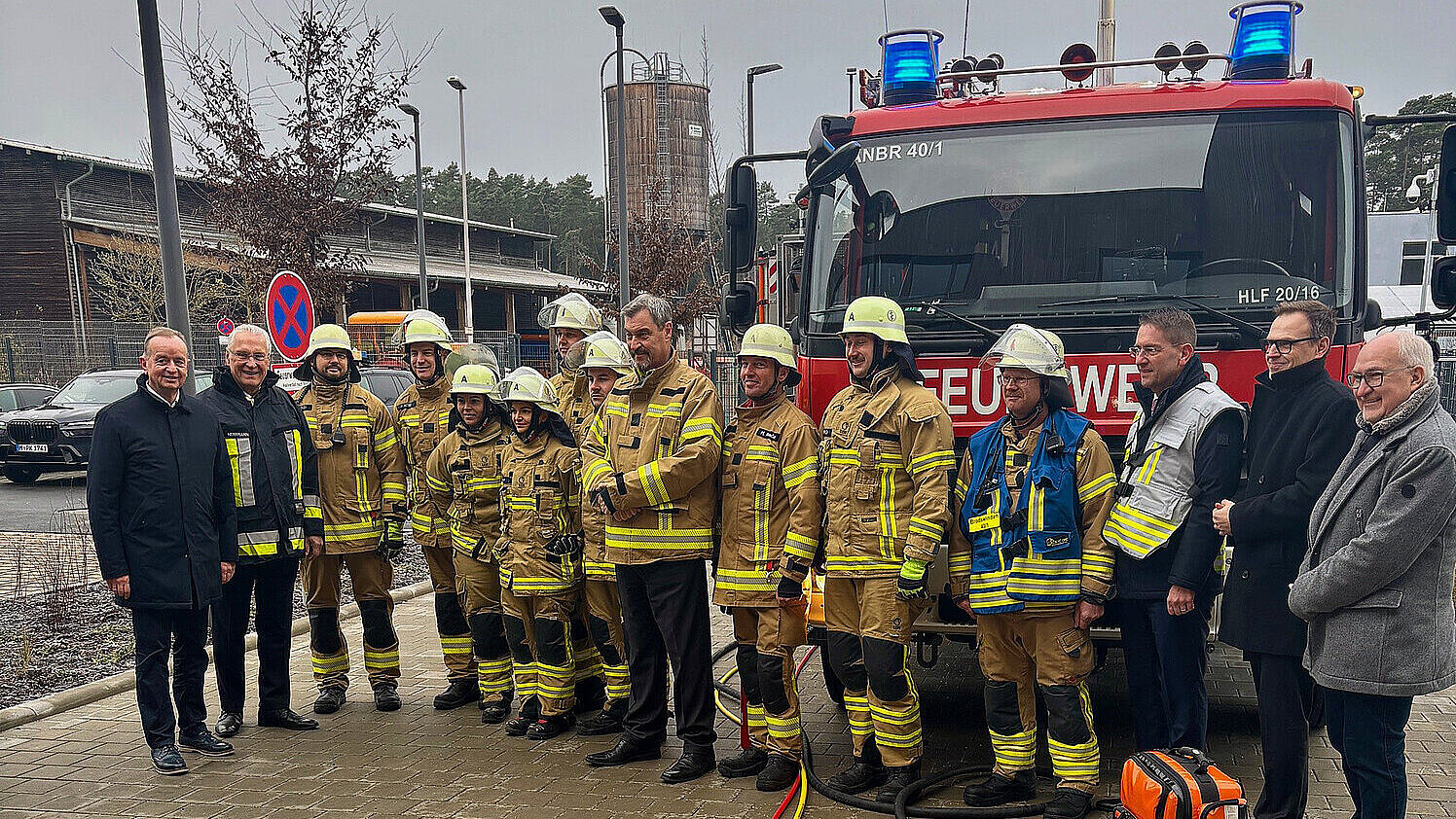 Eine Gruppe von Feuerwehrleuten in gelben Schutzanzügen und Helmen steht vor einem roten Feuerwehrfahrzeug mit Blaulicht. Vor ihnen sind verschiedene Feuerwehrgeräte und Ausrüstungsgegenstände auf einem roten Untergrund ausgelegt. Die Szene spielt sich draußen auf einem gepflasterten Platz ab.