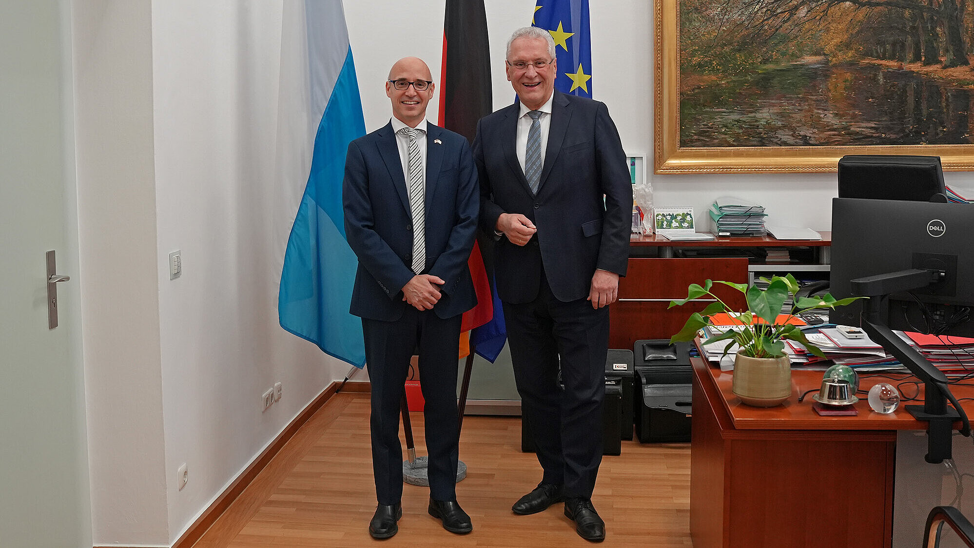 US-GK Dr. James Miller li. im Bild und Innenminister Joachim Herrmann re. im Bild nebeneinander in einem hellen Büro mit Holzfußboden. Hinter ihnen stehen drei Flaggen auf Ständern; rechts an der Wand hängt ein großes Gemälde. Im Vordergrund rechts ist ein Schreibtisch mit Computerbildschirm, Akten und einer Topfpflanze zu sehen. Die Gesichter wurden im Bild unkenntlich gemacht.