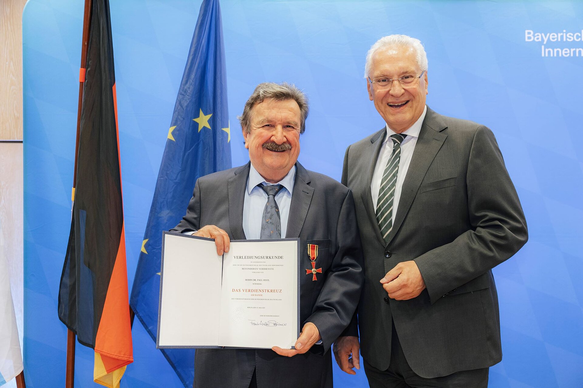 Auf dem Bild ist Innenminister Joachim Herrmann mit dem Geehrten Dr. Paul Festl zu sehen, hinter ihnen eine Pressewand sowie die bayerische, die deutsche und die EU-Fahne