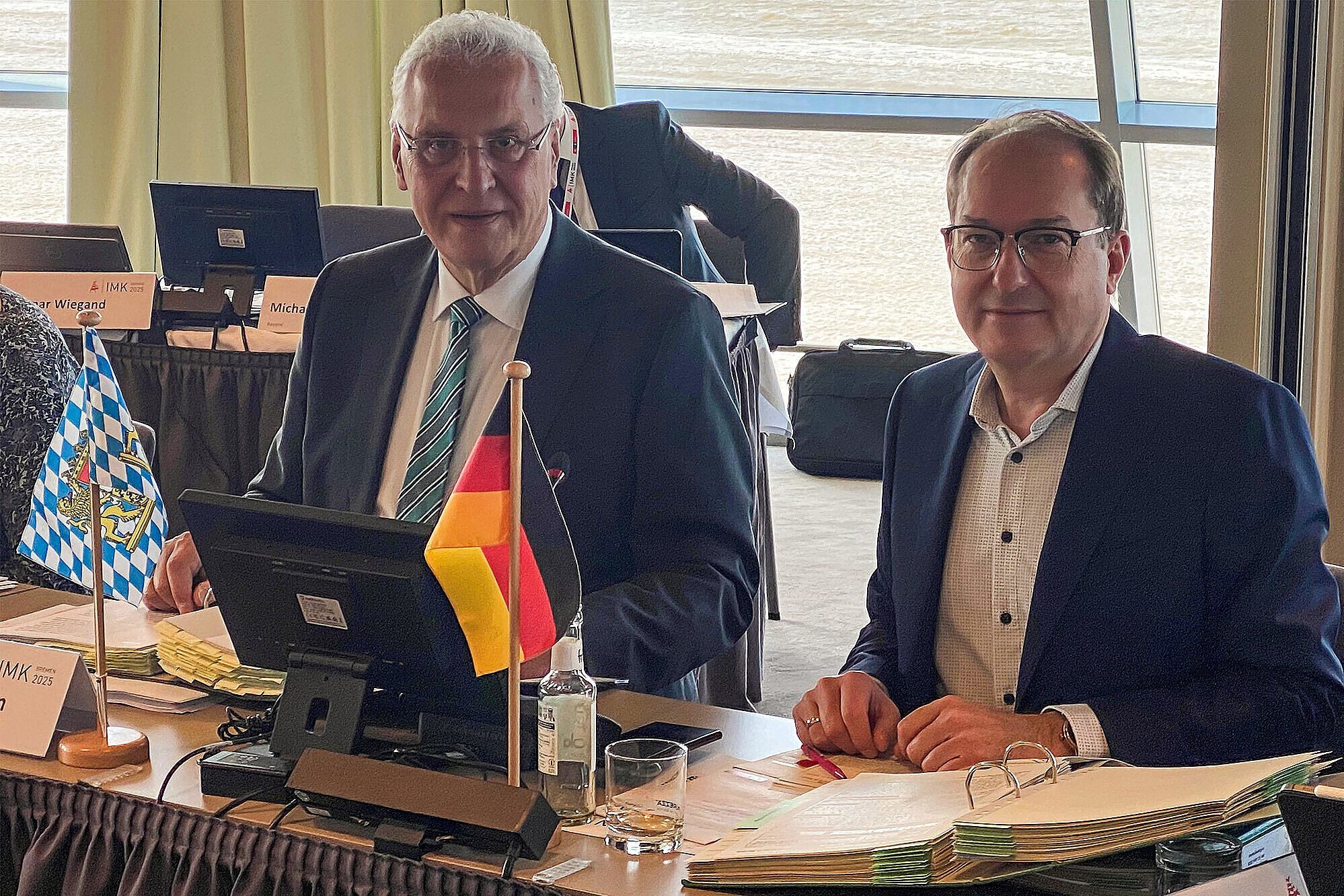 Zwei Männer im Anzug sitzen nebeneinander an einem Konferenztisch bei der Innenministerkonferenz 2025 in Bremerhaven. Vor ihnen stehen ein Bildschirm, ein Mikrofon, Ordner mit Dokumenten, eine Wasserflasche sowie zwei kleine Tischflaggen – die bayerische Rautenflagge und die deutsche Nationalflagge. Im Hintergrund sind weitere Teilnehmende an Tischen sowie große Fenster mit Blick auf eine Wasserfläche zu sehen.
