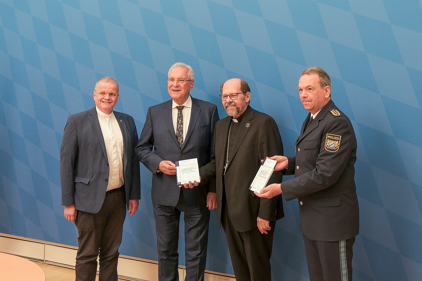 Gruppenbild von rechts Monsignore Simbeck, Innenminister Joachim Herrmann und ganz links LPP Michael Schwald vor der blauen Pressewand im Raum 111 im StMI