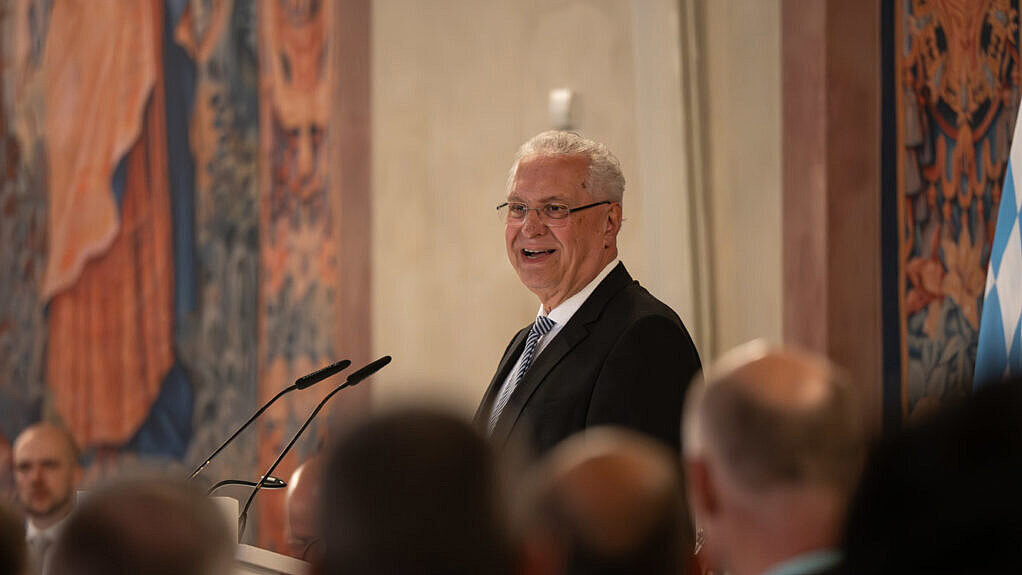 Redner in Anzug steht am Pult vor Publikum in festlichem Saal; Wandteppiche und Flagge im Hintergrund; Gesichter teils unkenntlich.