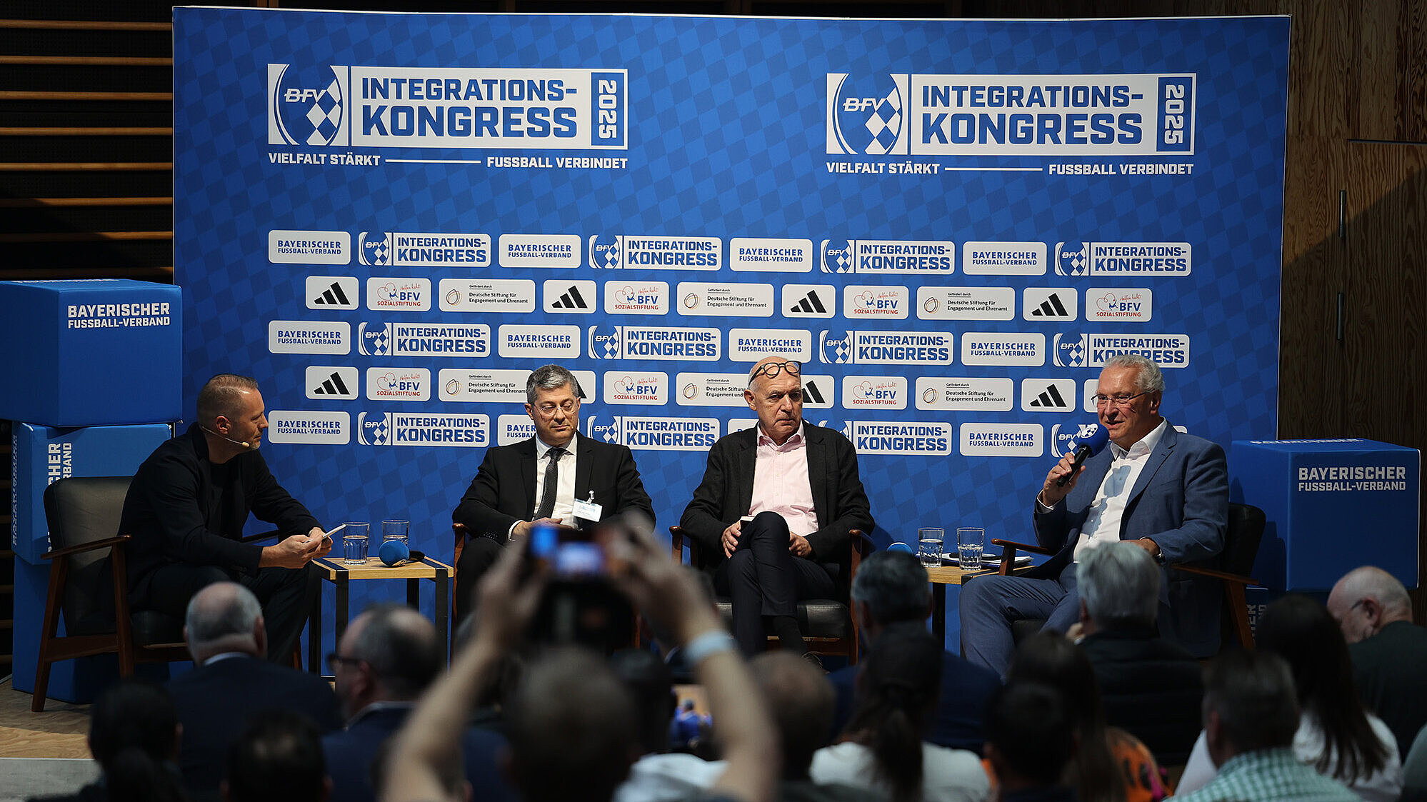 Auf dem Bild ist eine Podiumsdiskussion beim 1. BFV-Integrationskongress in Herzogenaurach zu sehen. Vier Männer sitzen auf der Bühne vor einem blauen Hintergrund mit dem Logo des Kongresses und verschiedenen Sponsoren. Ein Mann in einem blauen Anzug hält ein Mikrofon in der Hand und spricht, während die anderen drei Männer aufmerksam zuhören. Im Publikum sind zahlreiche Zuschauer zu sehen, die die Diskussion verfolgen.