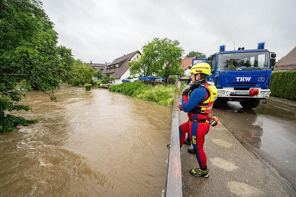 Ein DLRG Mitarbeiter steht auf einer Brücke und schaut auf reißendes Hochwasser herab, im Hintergrund steht ein THW-Fahrzeug