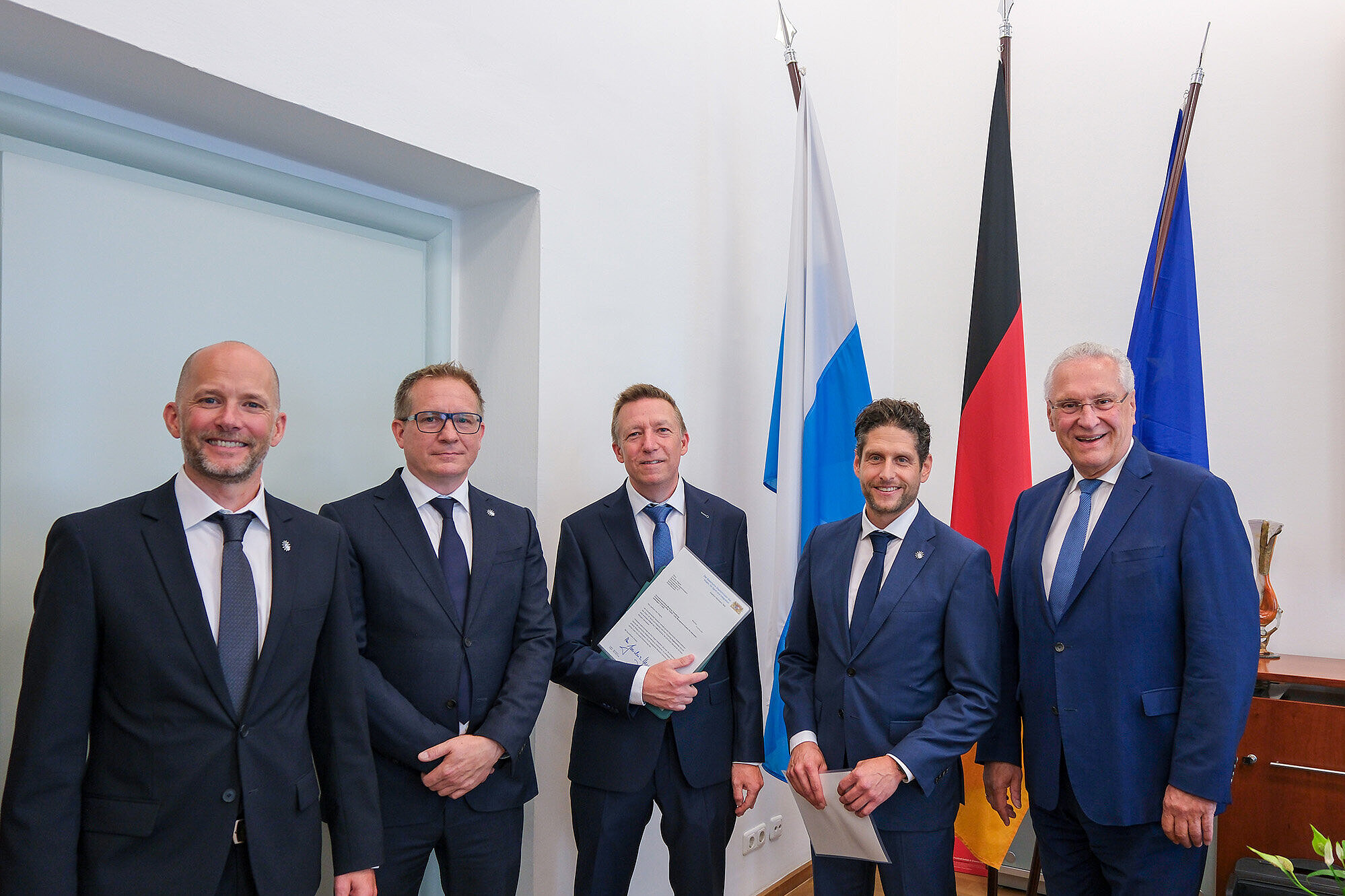 Fünf Männer in dunklen Anzügen stehen lächelnd nebeneinander in einem offiziellen Raum. Zwei von ihnen halten Urkunden in der Hand. Im Hintergrund stehen die bayerische, deutsche und europäische Flagge. Es handelt sich um die Übergabe einer Prämierungsurkunde für einen Verbesserungsvorschlag.