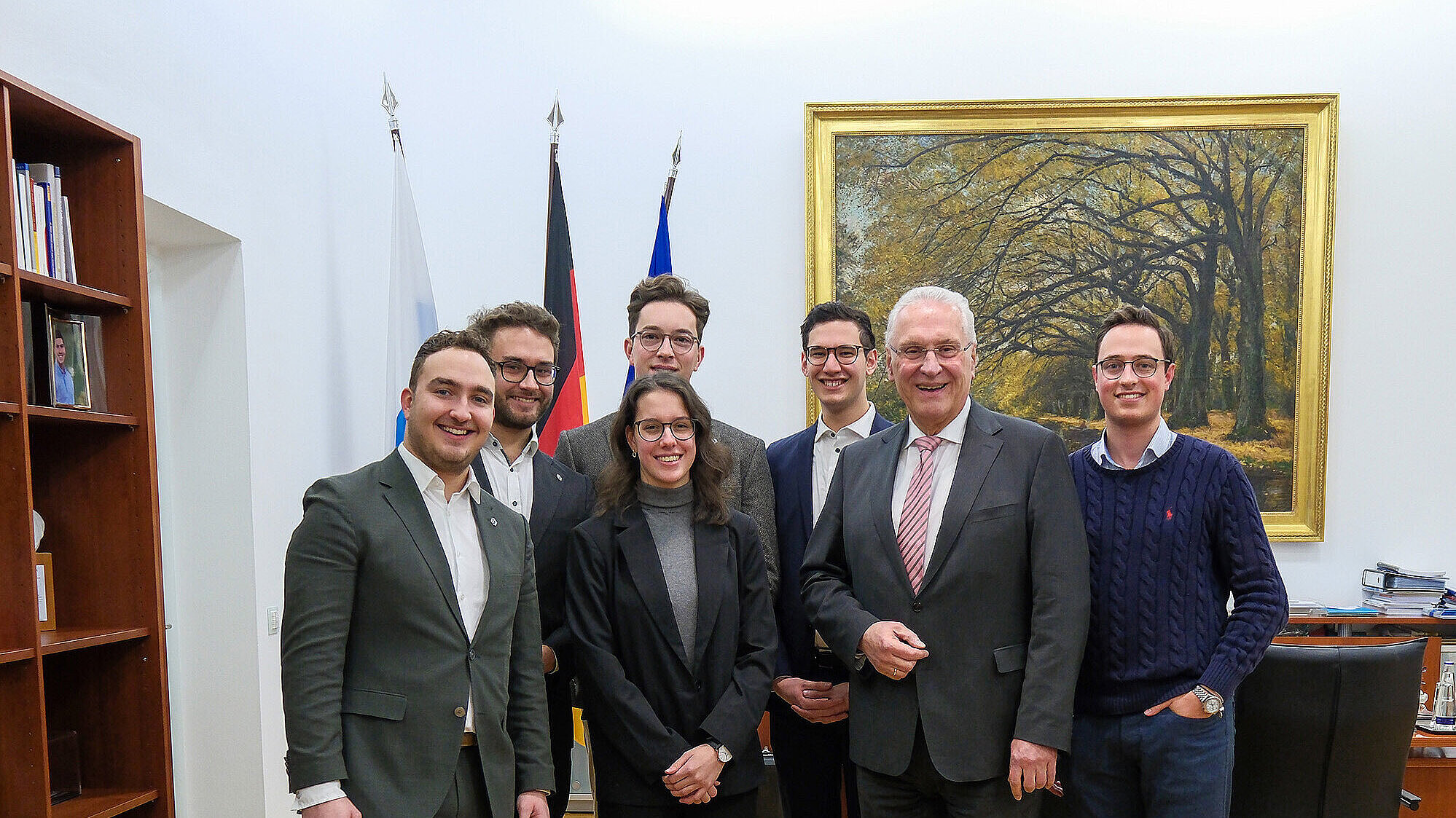 Eine Gruppe von 7 Personen (Landesvorstand des RCDS Bayern e.V., 2. Von rechts Innenminister Joachim Herrmann) steht in einem Büro mit Holzboden. Im Hintergrund sind drei Fahnen zu sehen: die Bayerische, die Deutsche und die Europäische Flagge. An der Wand hängt ein großes Bild mit einem herbstlichen Waldweg. Rechts im Bild ist ein Schreibtisch mit Stühlen. Die Personen tragen formelle und semi-formelle Kleidung.
