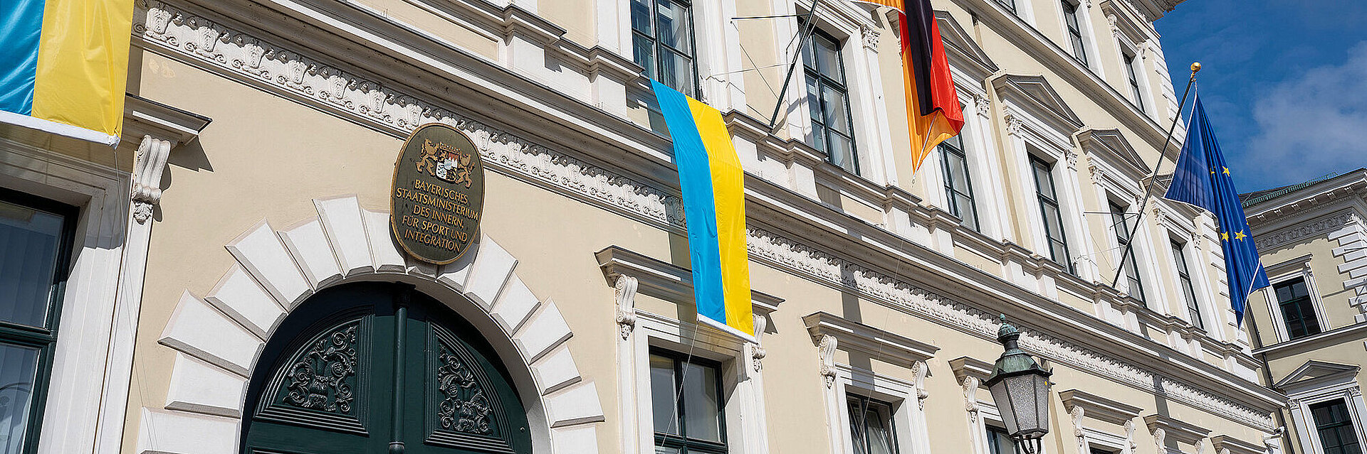 Neoklassizistische Fassade eines Amtsgebäudes mit verziertem dunkelgrünen Eingangstor, mehreren Fenstern, zwei hängenden ukrainischen Flaggen, deutscher und EU‑Flagge sowie Straßenlaterne und Gehweg im Vordergrund