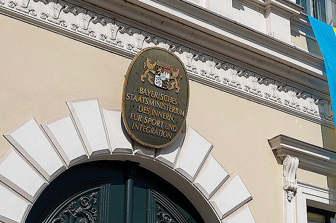 Neoklassizistische Fassade eines Amtsgebäudes mit verziertem dunkelgrünen Eingangstor, mehreren Fenstern, zwei hängenden ukrainischen Flaggen, deutscher und EU‑Flagge sowie Straßenlaterne und Gehweg im Vordergrund