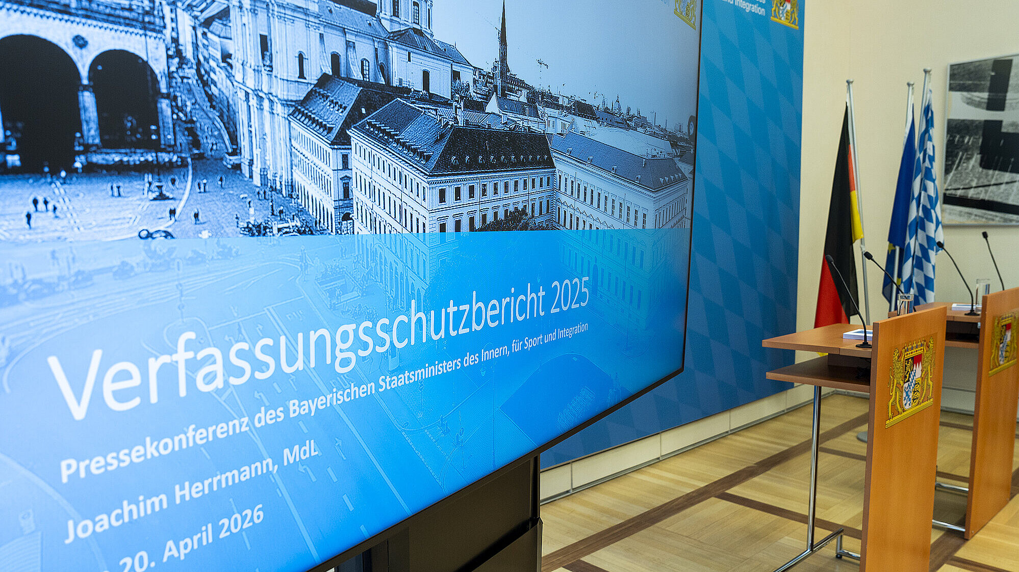 Auf dem Bild sieht man die Bildschirmpräsentation zur Pressekonferenz, dahinter die beiden (noch leeren) Rednerpulte.