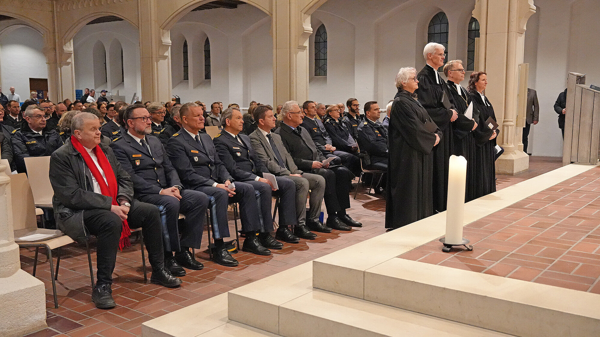 Eine Gruppe von Menschen sitzt in einer Kirche auf Stühlen, darunter mehrere Personen in Polizeiuniformen und einige in Anzügen. Vor ihnen stehen vier Geistliche in schwarzen liturgischen Gewändern. Im Vordergrund steht eine brennende weiße Kerze auf einem erhöhten Altarbereich aus Stein und Fliesen. Die Kirche hat hohe, gewölbte Fenster und Säulen.
