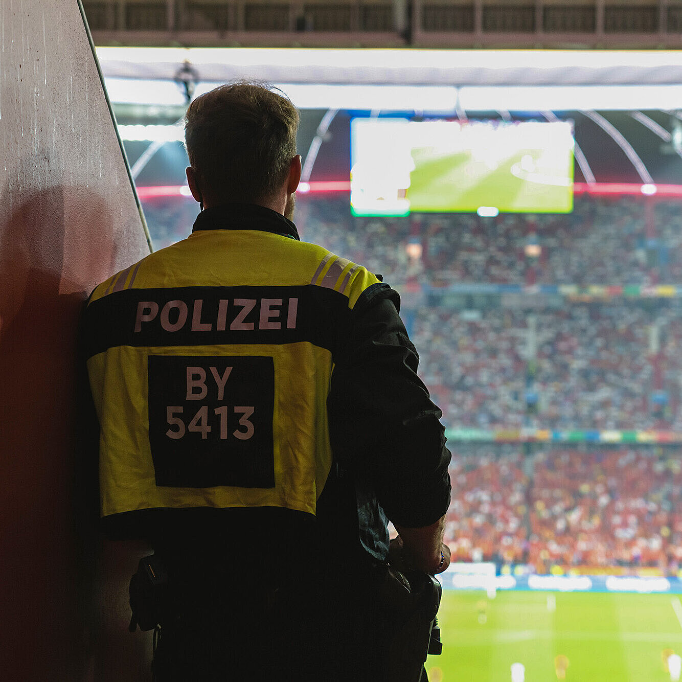 Ein Polizist steht in einem Stadion, das für ein Fußballspiel vorbereitet ist. Er trägt eine gelbe Weste mit der Aufschrift „POLIZEI“ und einer Nummer. Im Hintergrund sind viele Zuschauer im Stadion zu sehen, sowie eine große Anzeigetafel.