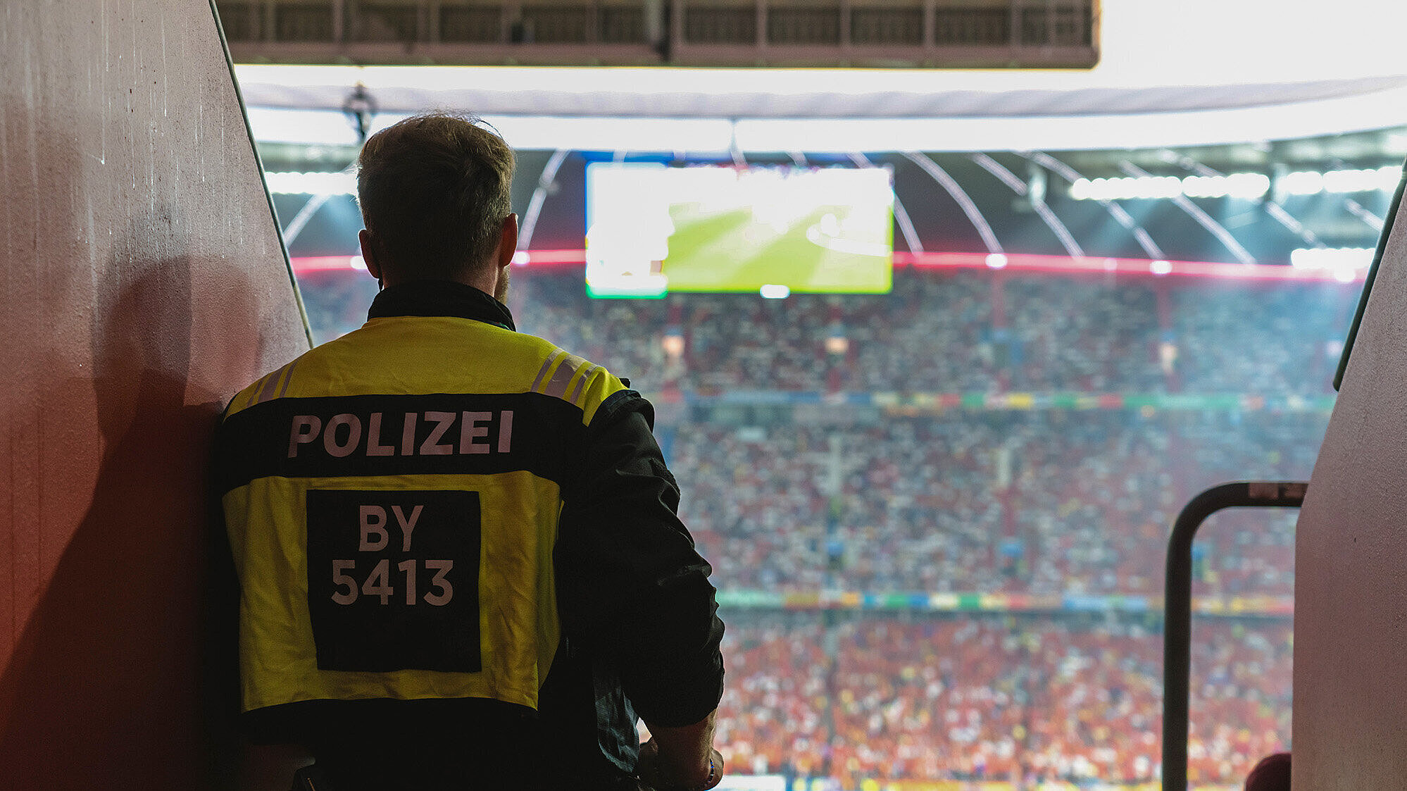 Ein Polizist steht in einem Stadion, das für ein Fußballspiel vorbereitet ist. Er trägt eine gelbe Weste mit der Aufschrift „POLIZEI“ und einer Nummer. Im Hintergrund sind viele Zuschauer im Stadion zu sehen, sowie eine große Anzeigetafel.